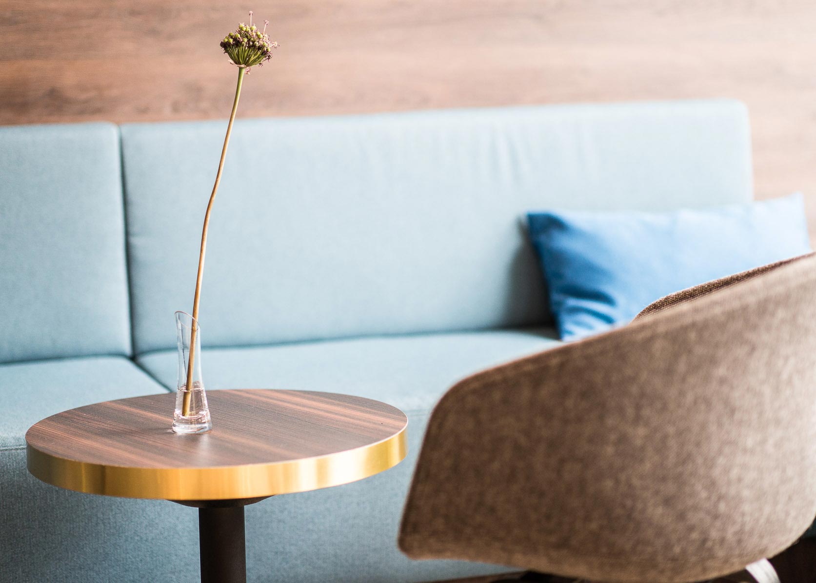 Bild von einem braunen runden Holztische mit einer kleinen Vase und Blume in der Mitte des Tisches. Im Hintergrund ist ein blauer sofa mit einem dunkleren blauen kissen zu und einen braunen stoff stuhl mit lehne zu sehen. 

