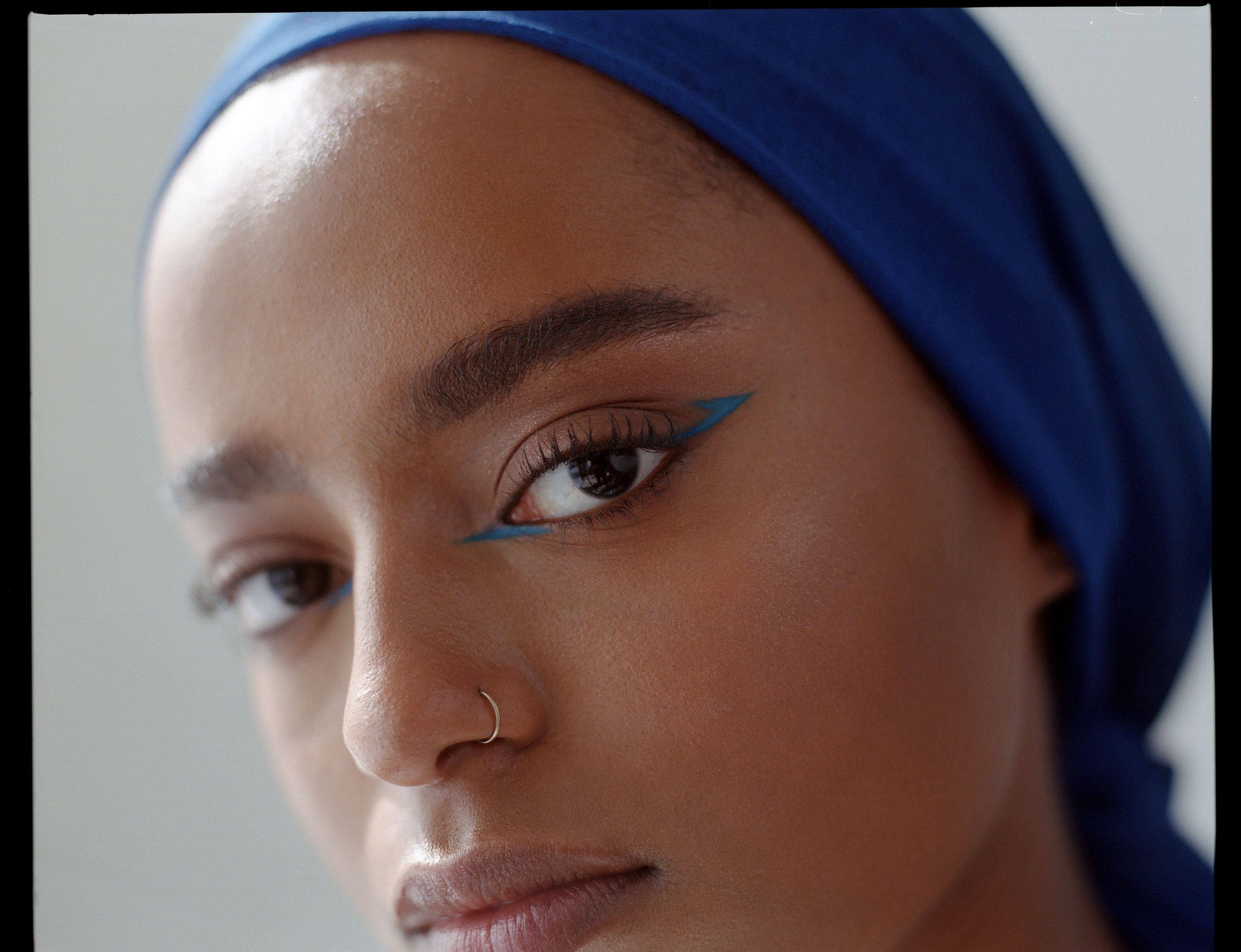 Portrait of a Black woman with turquoise eye shadow, gold nose ring and blue headscarf looking obliquely at the camera from the left, shot by Samia Rachel

