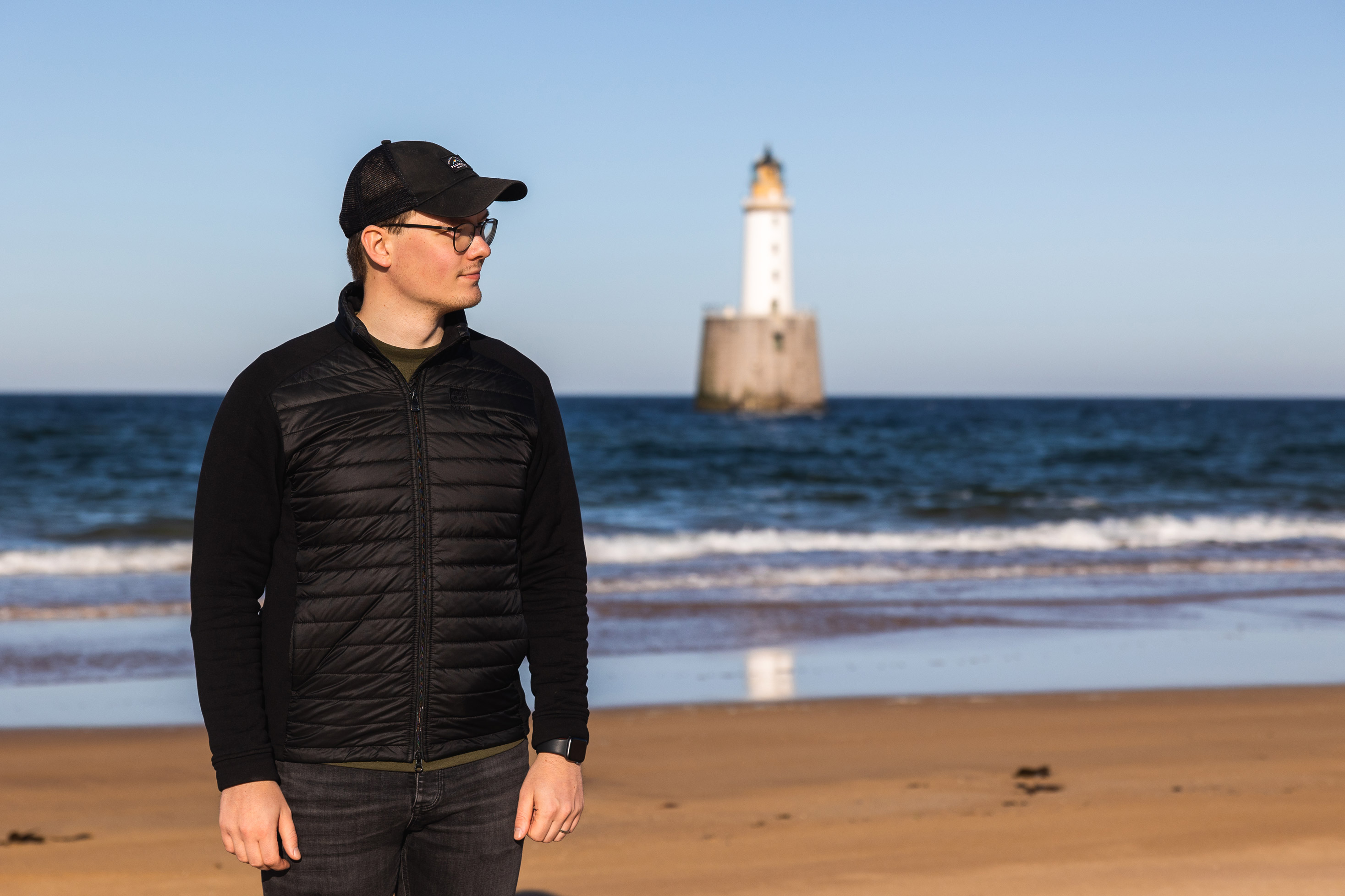 a man wearing a hat standing on a beach with the sea and a lighthouse in the background