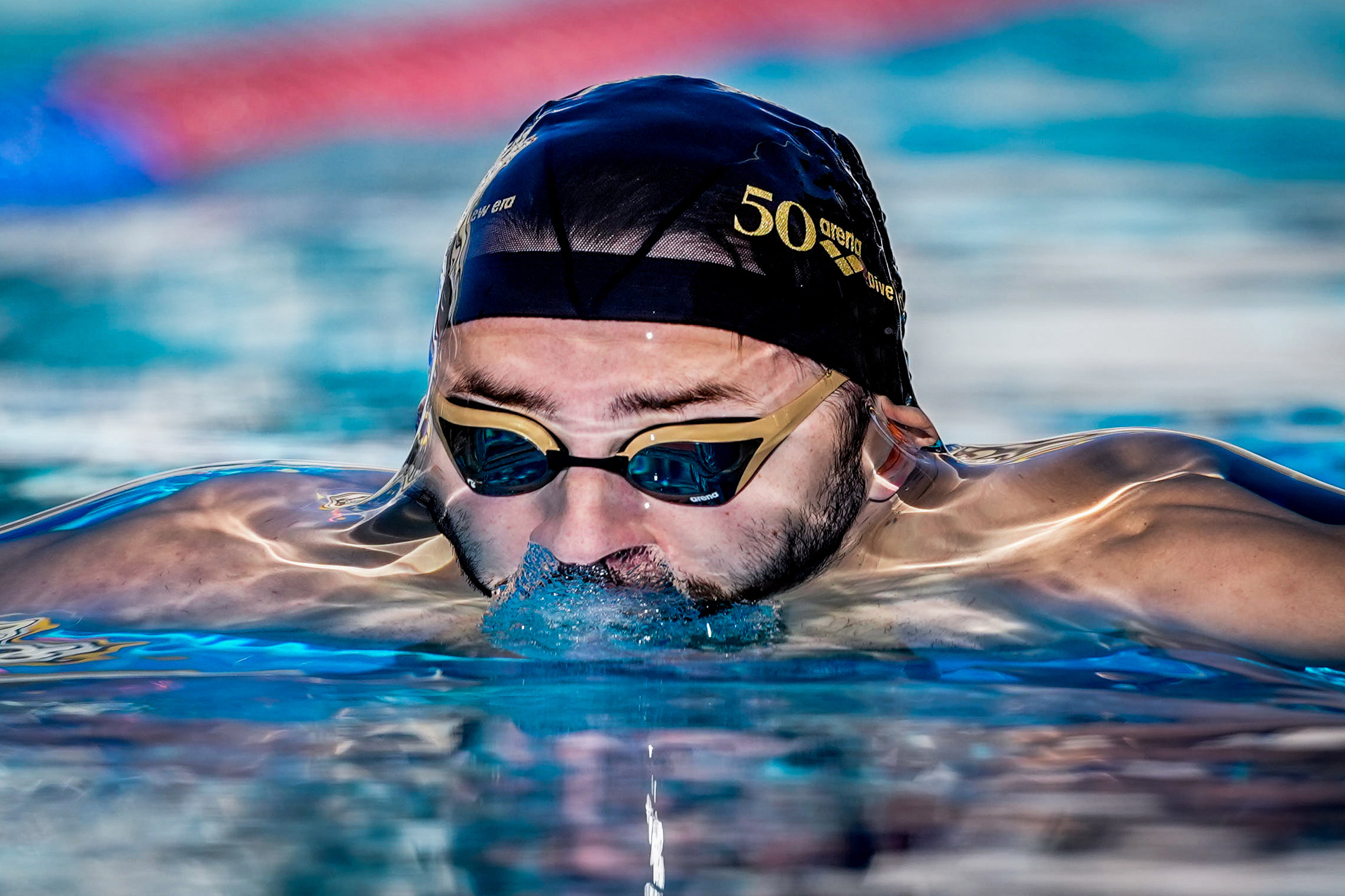À courte distance, un nageur portant un bonnet de natation noir et des lunettes de protection émerge de l'eau sans rompre la surface.

