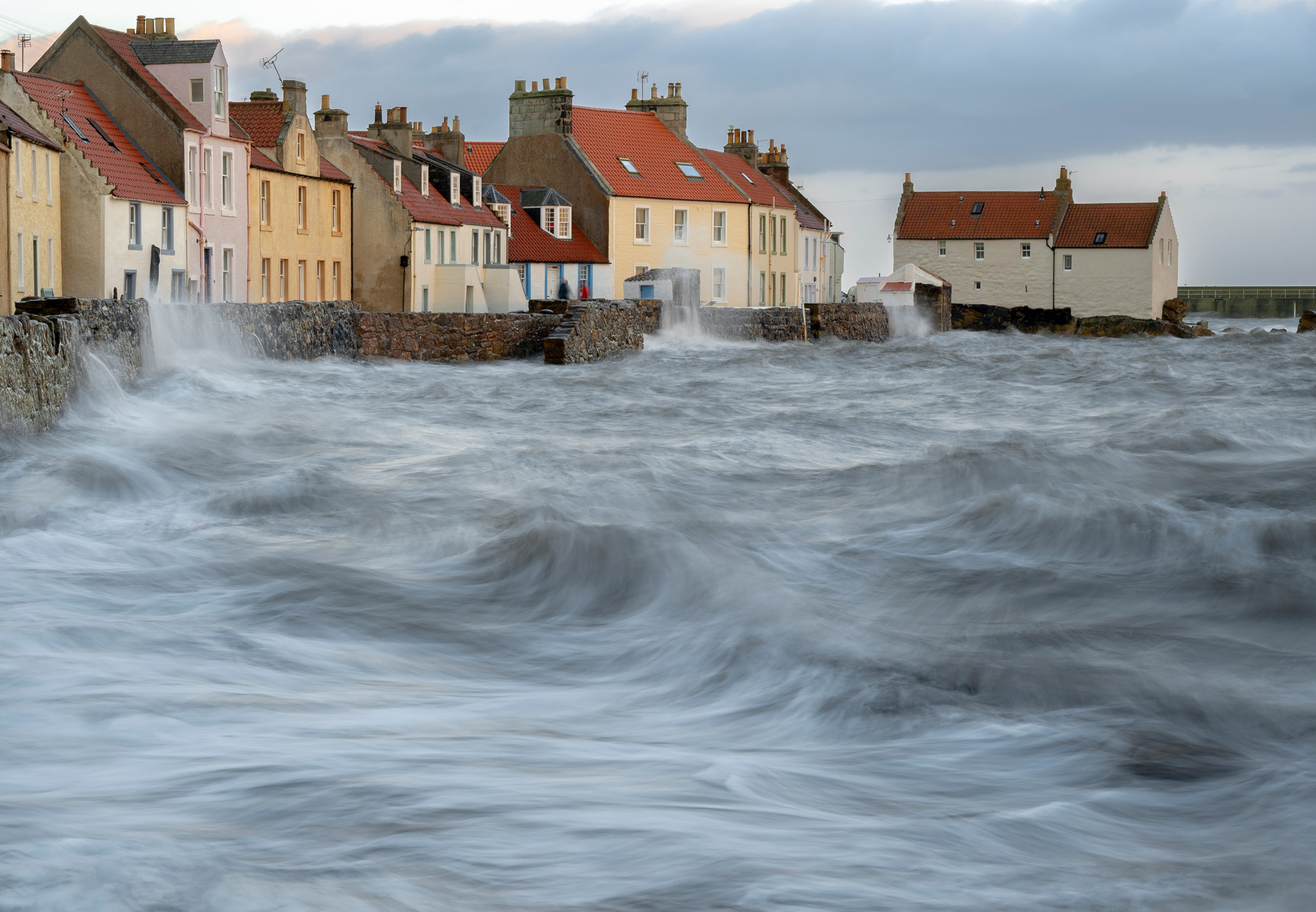 Waves crash into a wall below a row of houses by the coast in Pittenweem