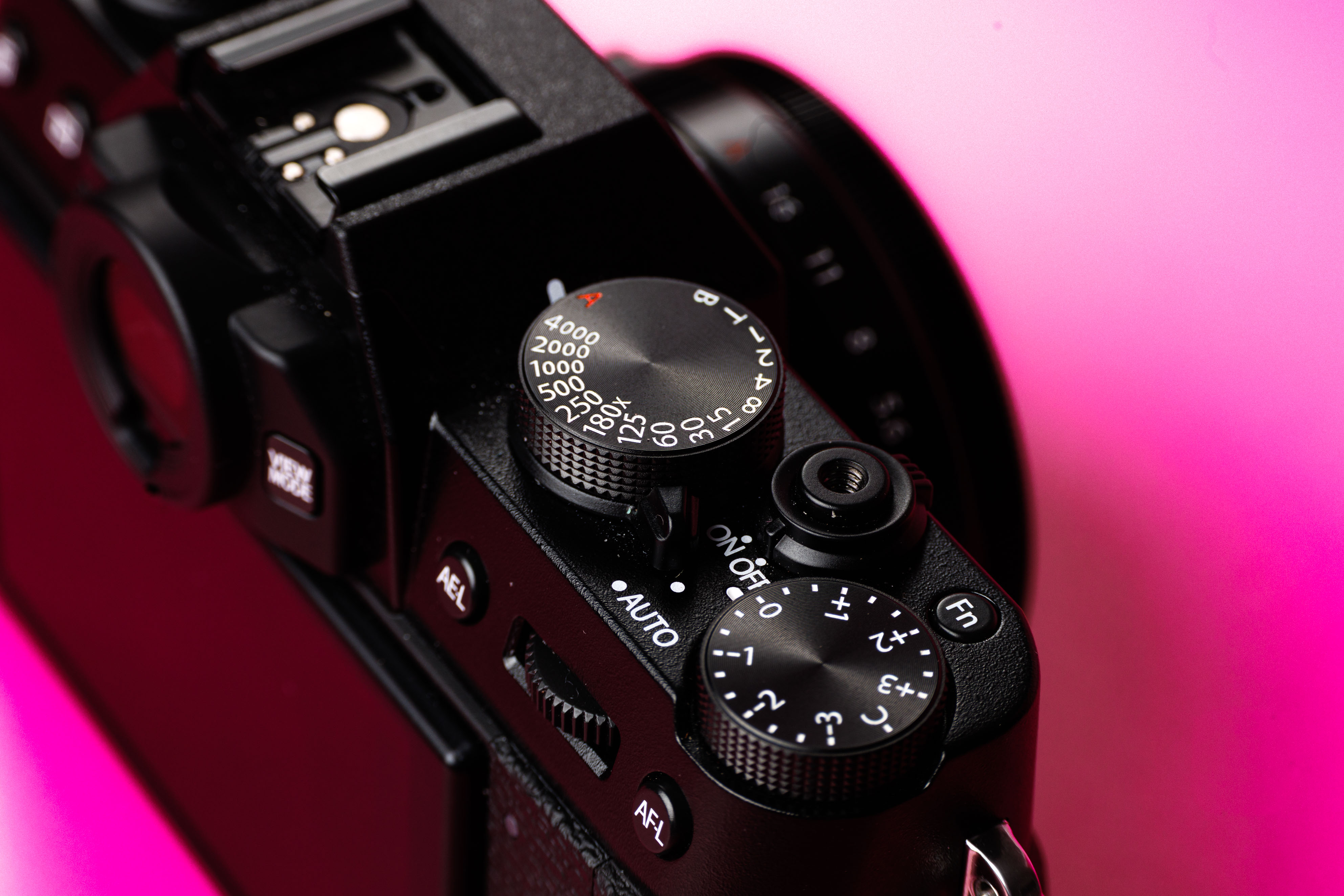 Close-up of Fujifilm X-T30 top plate showing shutter speed dial, exposure compensation and control layout against a vibrant pink background.