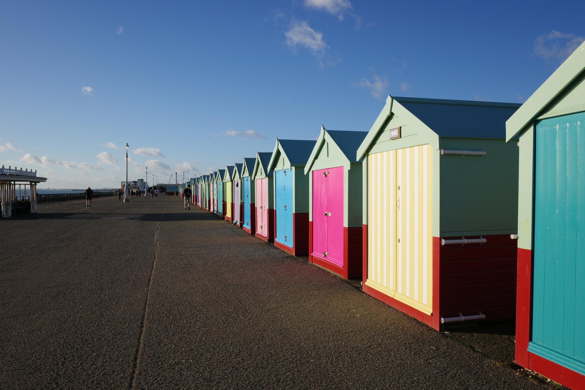 Een boulevard op een zonnige dag, vol met kleurrijke strandhuisjes. Boven je is een helderblauwe lucht met dunne witte wolken. Gemaakt met het Vivid-filmsimulatiefilter.