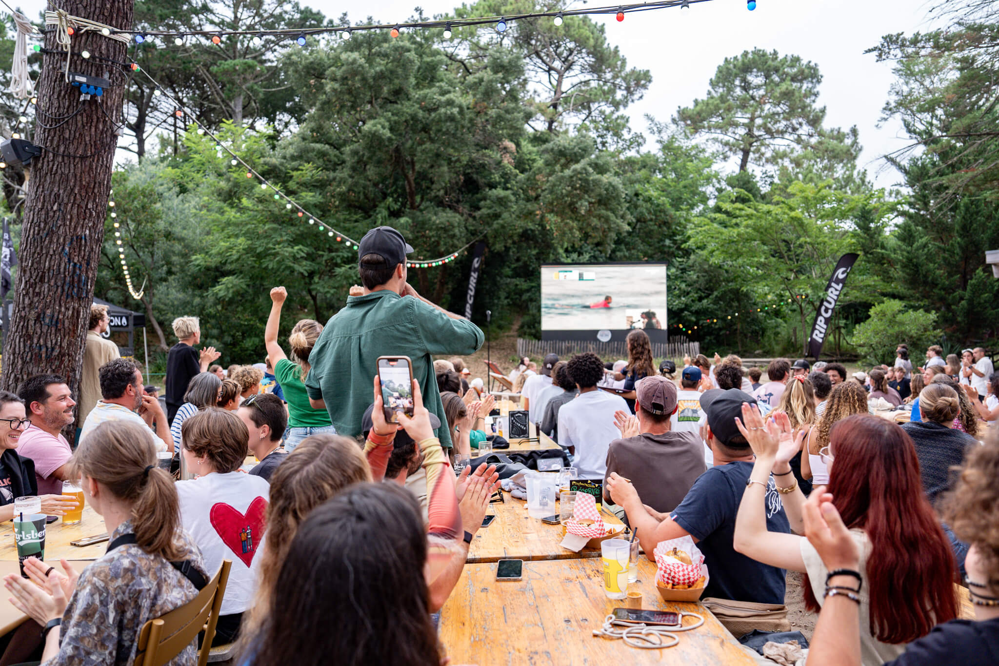 
Des festivaliers sont rassemblés autour des tables et regardent la compétition de surf. Certains filment avec leur téléphone et d’autres applaudissent. Photo par Paul Lavoine prise avec le Sony A9 III.

