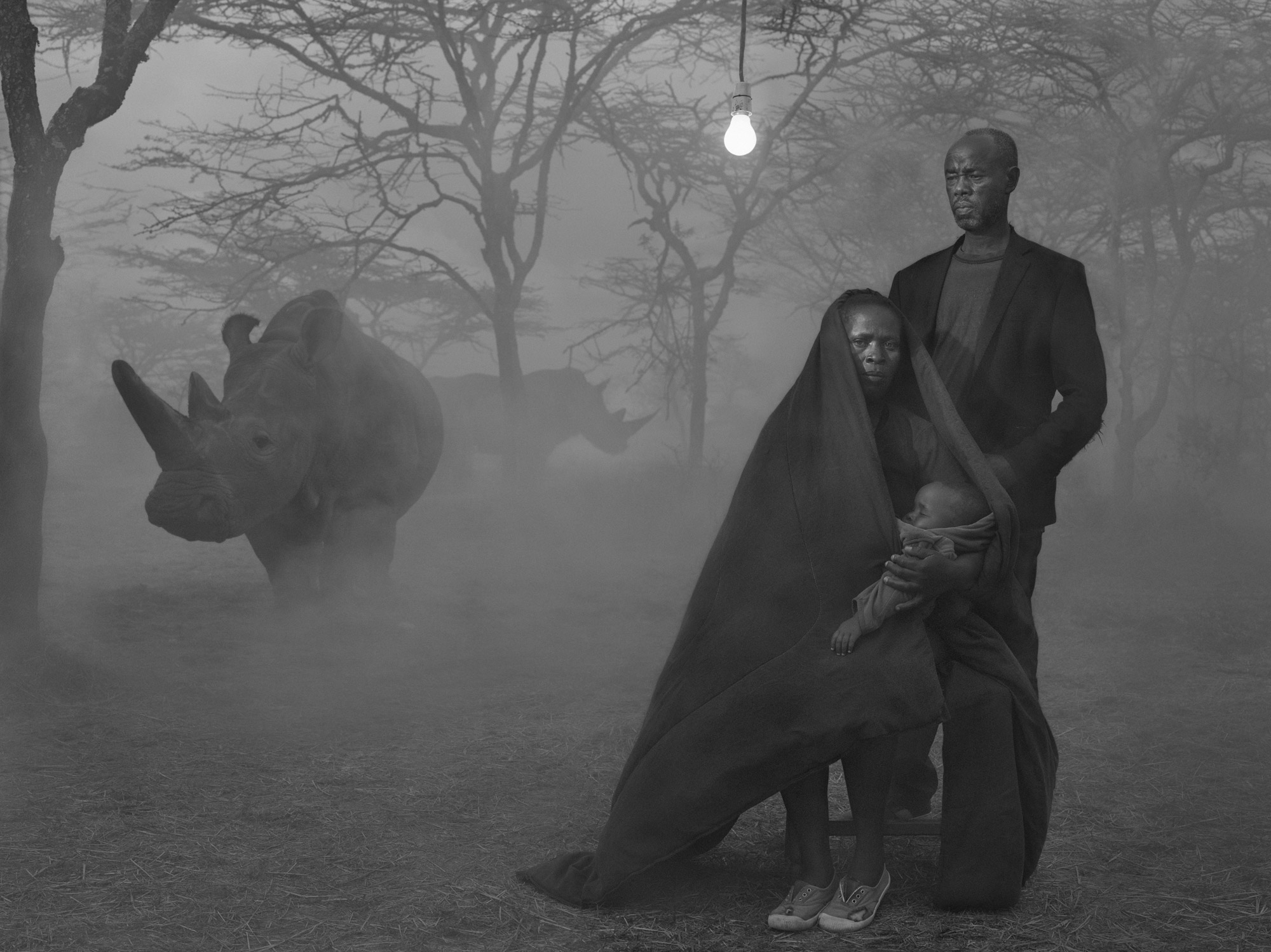 Ein Schwarz-Weiß-Bild einer Familie im Wald, umgeben von zwei Nashörnern im Hintergrund
