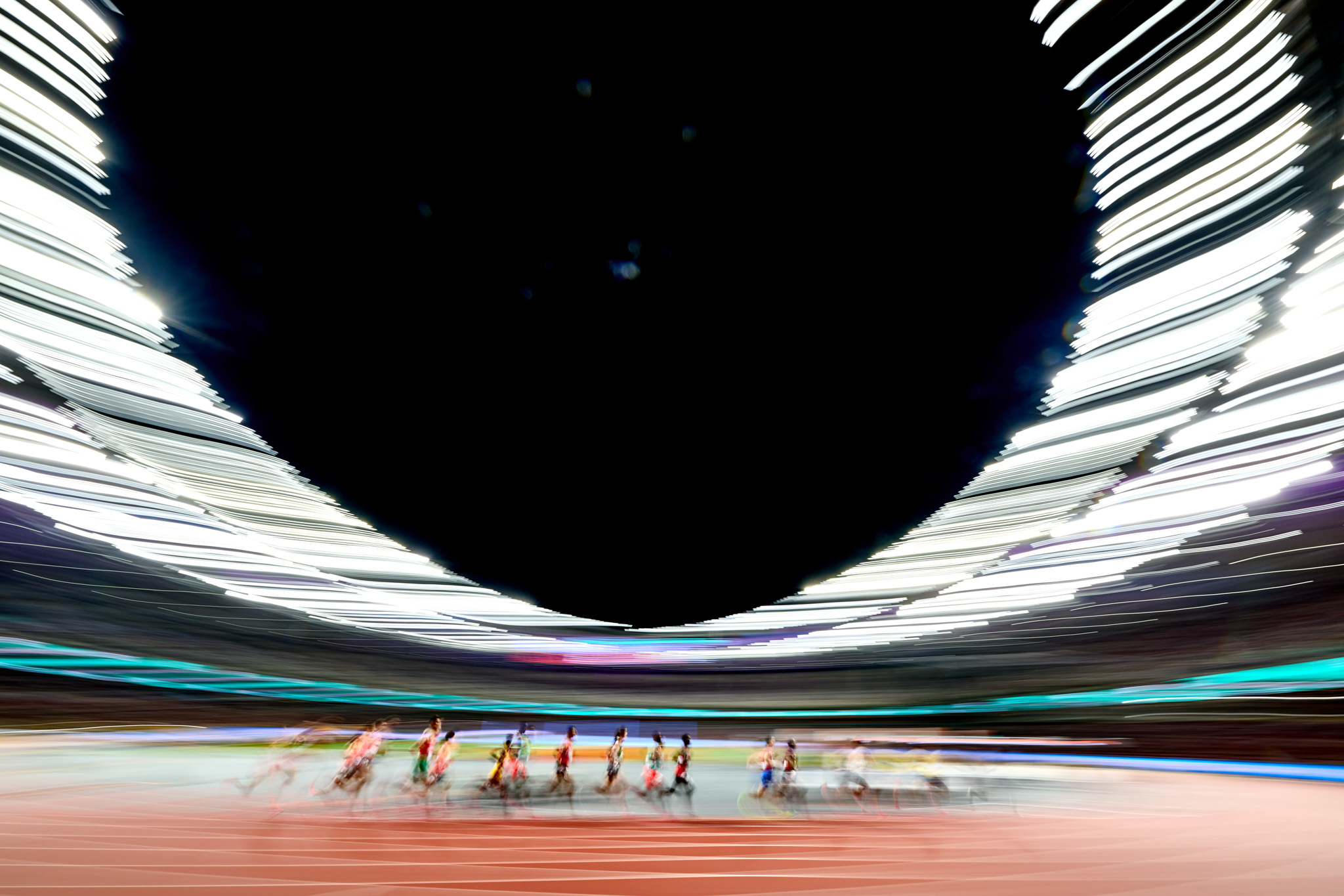 A wide shot of an athletics stadium as a group of athletes run past, who are blurry, creating a sense of motion. By Mattia Ozbot.