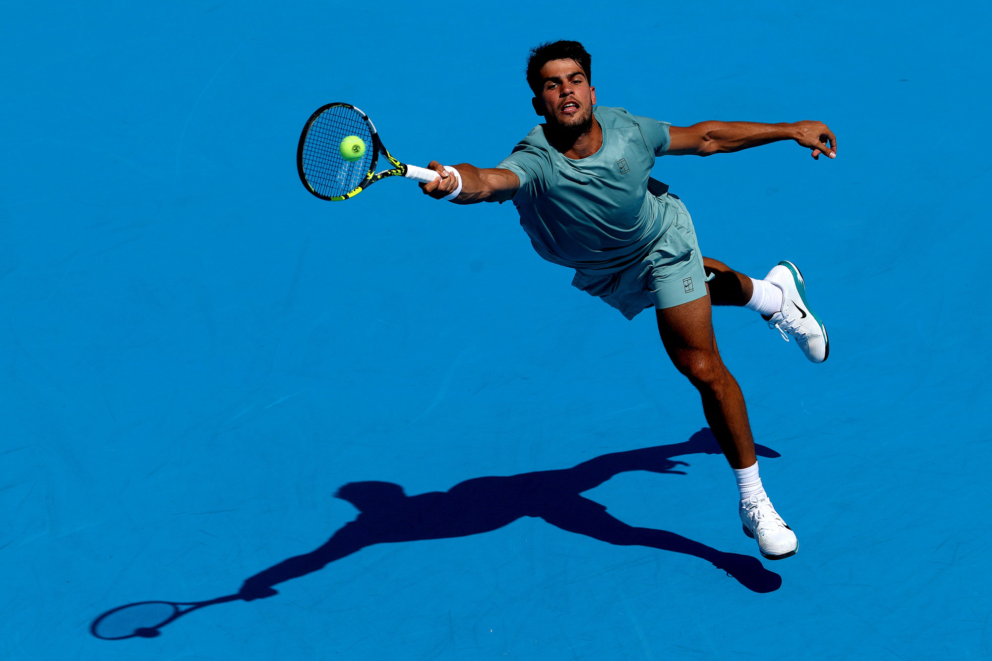 Carlos Alcaraz of Spain stretches for a ball while playing Hamad Medjedovic of Serbia during the Cincinnati Open in Mason, Ohio. His shadow is underneath him on a plain blue background.