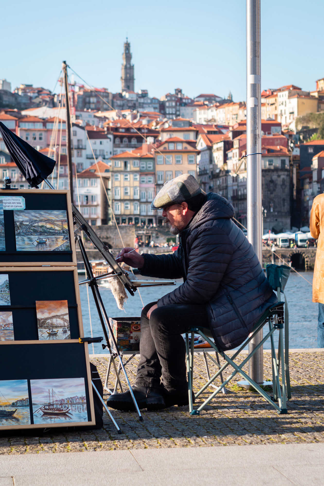 Person mit Mantel und Hut, die an der belebten Küste Portugals an einem sonnigen Tag auf einem Klappstuhl sitzt und malt.
