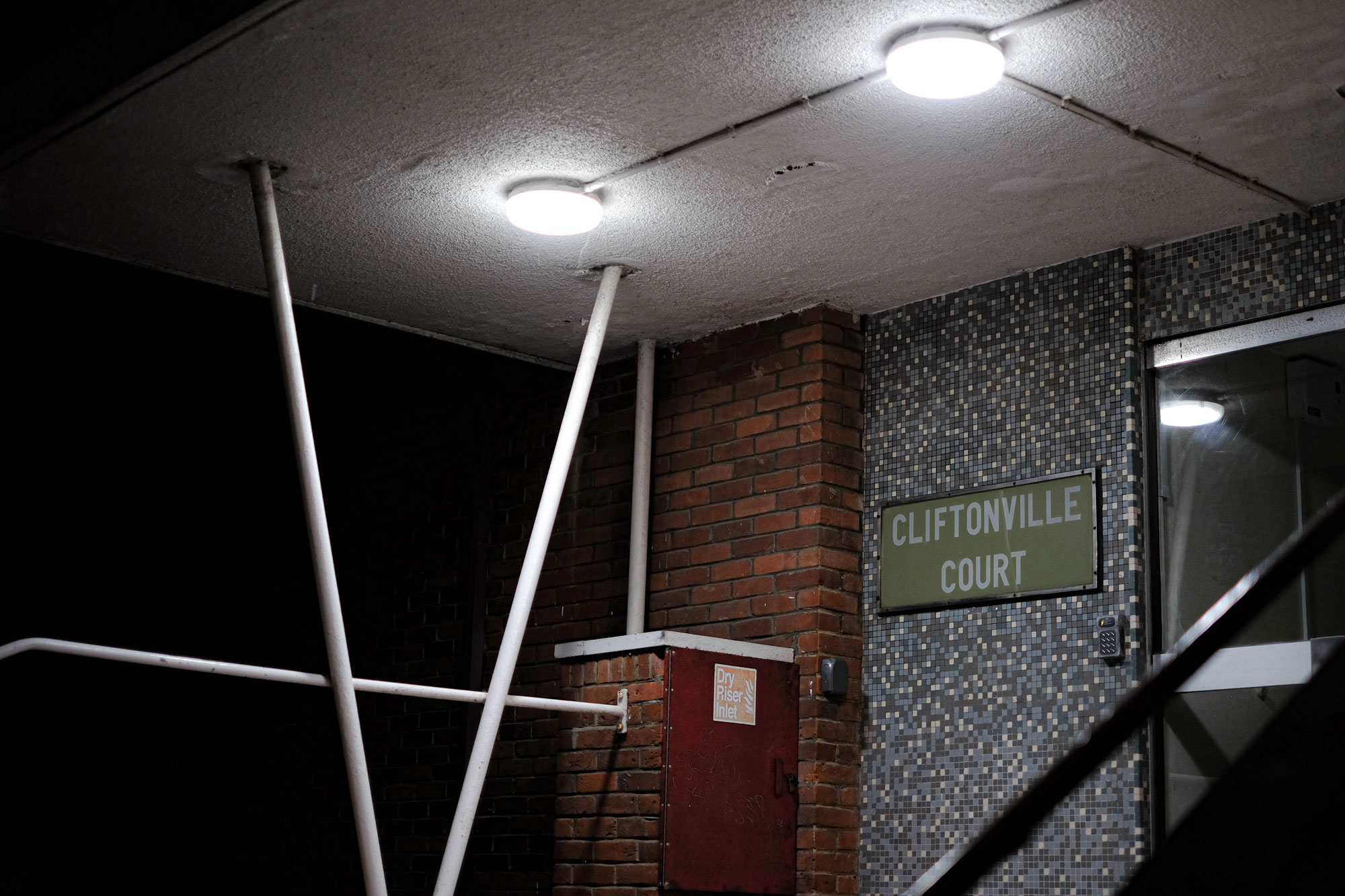 The entrance to a purpose-built block with a sign that reads ‘Cliftonville Court’. Two lights are on above the doorway, illuminating the red brick and mosaic details of the building.