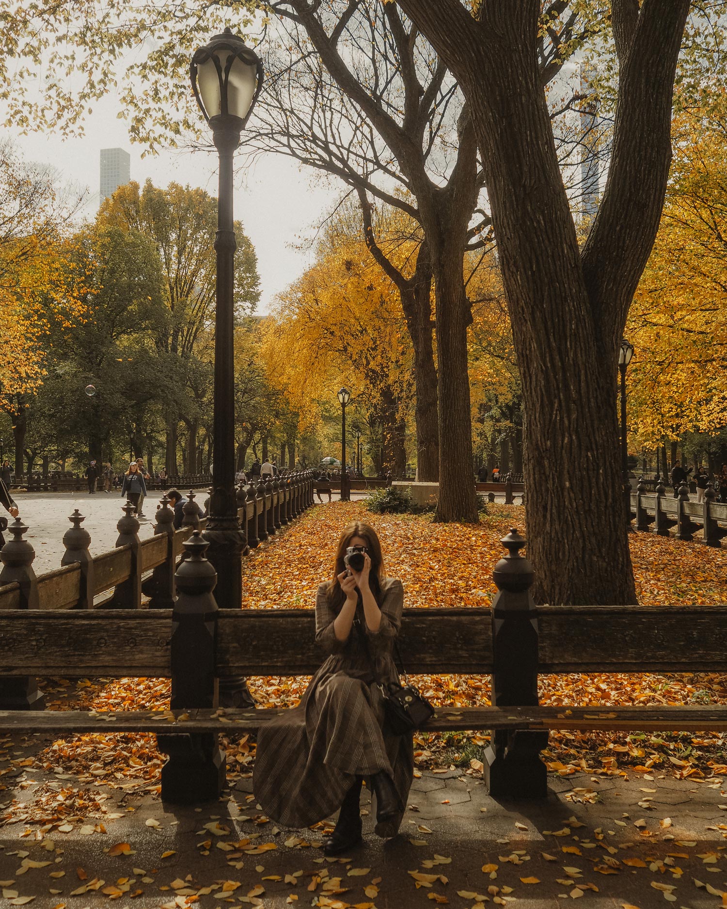 Georgina Lamrock holding a camera, sitting on a bench in Central Park, there are trees, buildings and a lamppost in the background. Shot on an Olympus PEN F by Georgina Lamrock