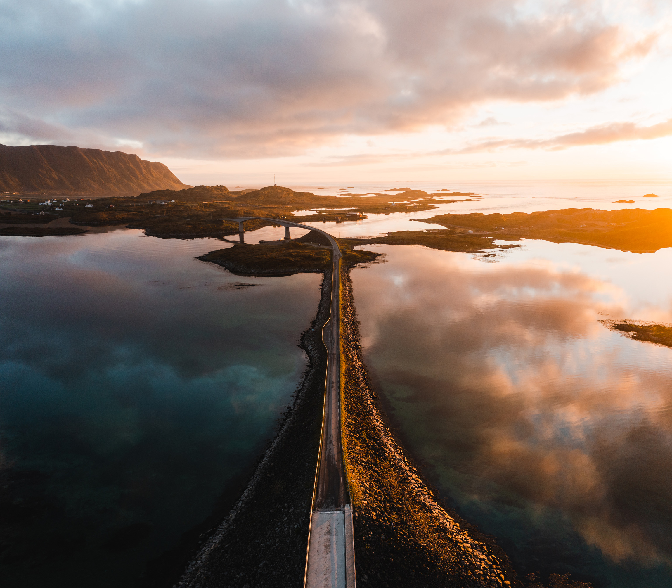 Bridge in Norway over water and small islands in Norway, the water is reflecting the orange sky and grey clouds, shot by Jordan Lancaster-Graham with a DJI Mavic II Pro