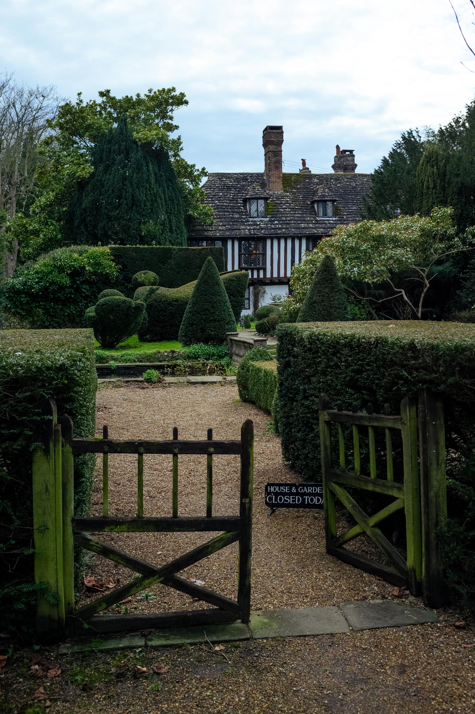 A house from the Tudor period peeks out from behind a well-manicured garden and trees. Two wooden gates are at the entrance of the garden, only one of them open.