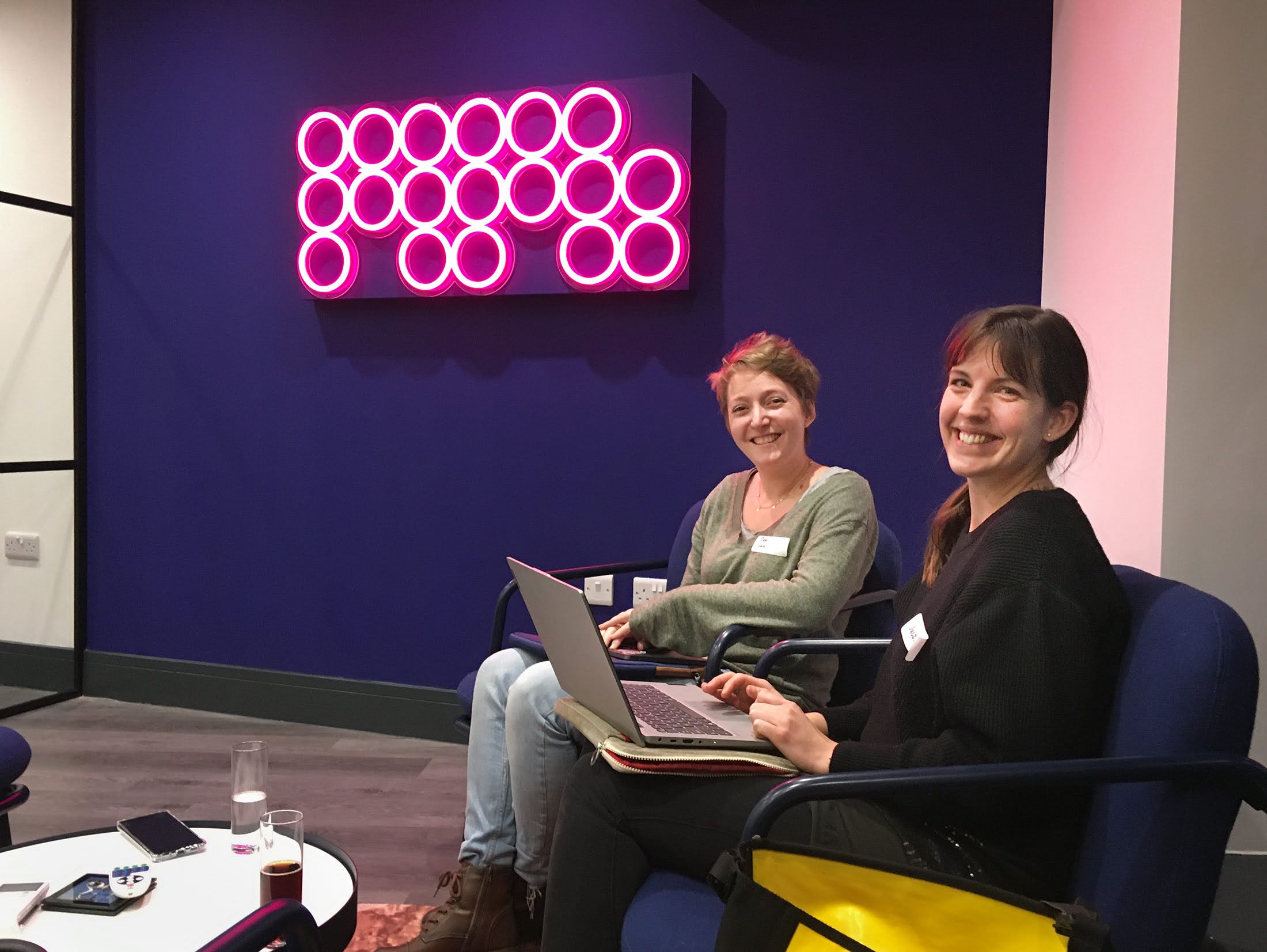 Two people sitting in chairs with laptops attending an MPB Brighton codebar event smiling at the camera. 