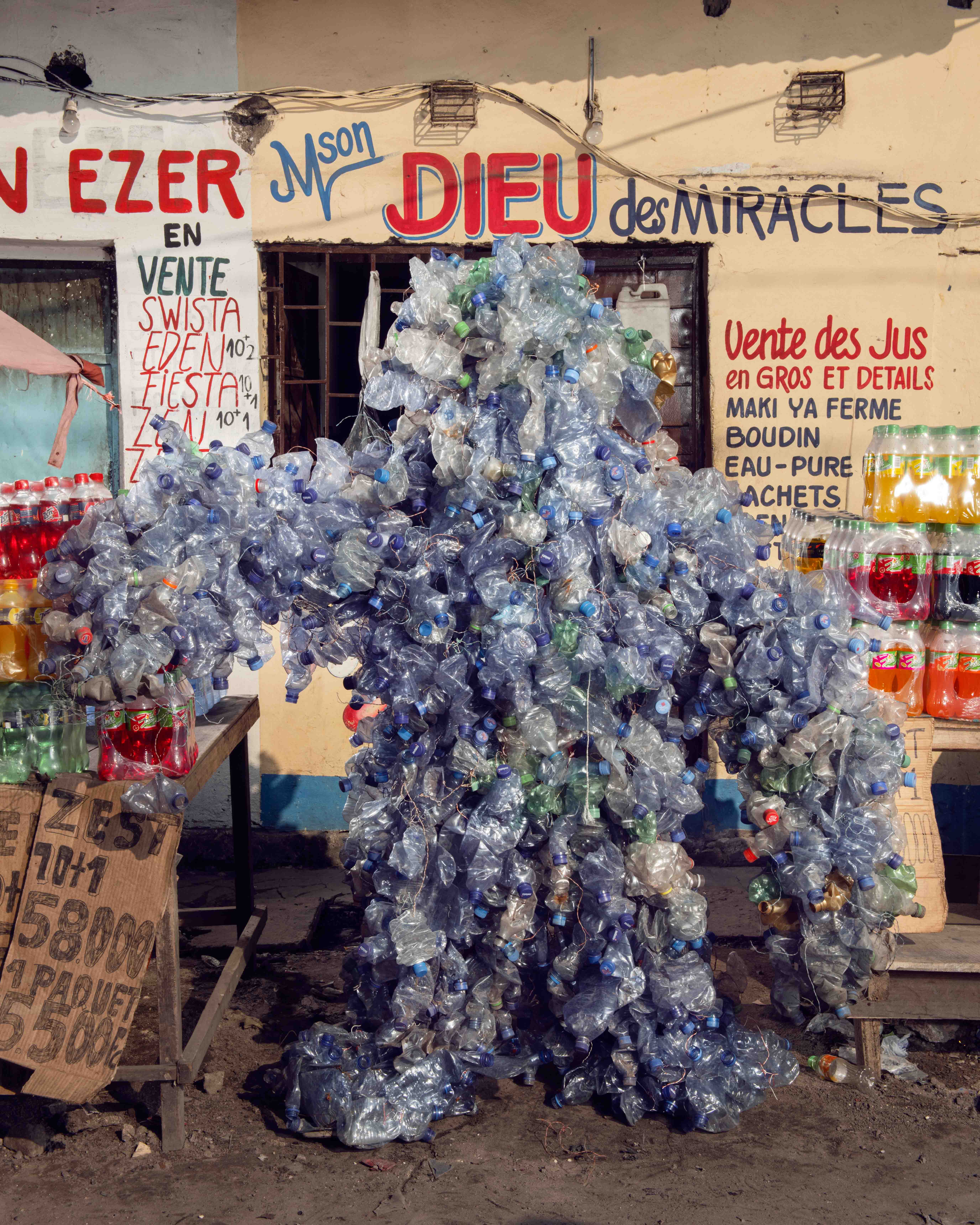 Wrapped in used plastic bottles, this Homo Detritus figure appears outside a juice shop in Kinshasa — a divine satire on waste and devotion.