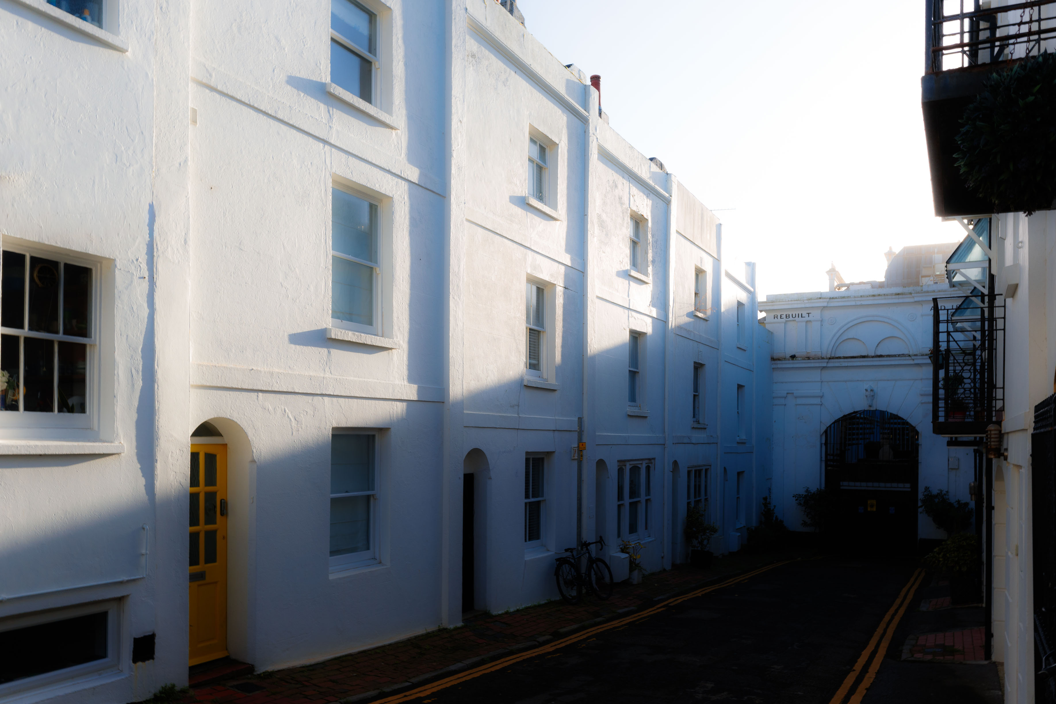Eine ruhige Straße mit weiß gestrichenen Gebäuden und einer gelben Haustür im weichen Abendlicht. Die tiefstehende Sonne wirft lange Schatten. Aufgenommen mit einer Ricoh GR IV.