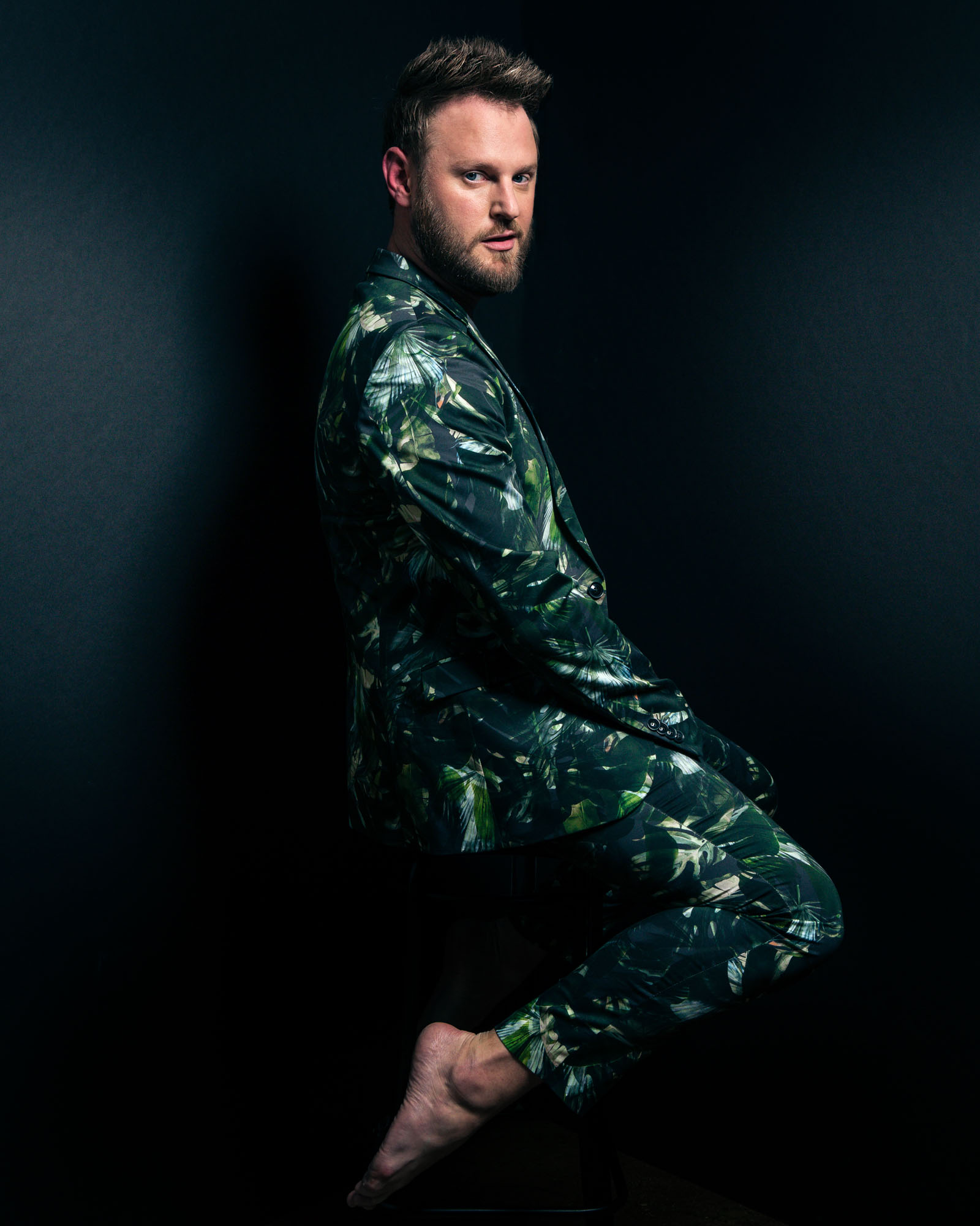 Bobby Berk, from Netflix series Queer Eye, wearing a green suit sitting barefoot on a stool, looking directly at the camera