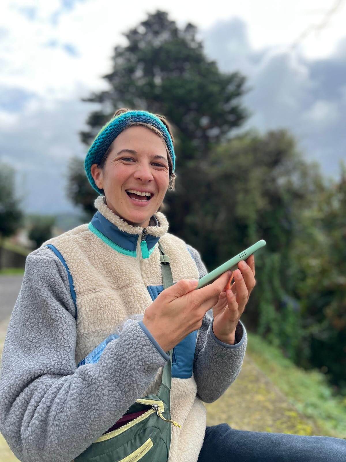 Eine warm gekleidete Person, die mit einem Telefon in der Hand in die Kamera lacht, mit einer bewaldeten Landschaft im Hintergrund.