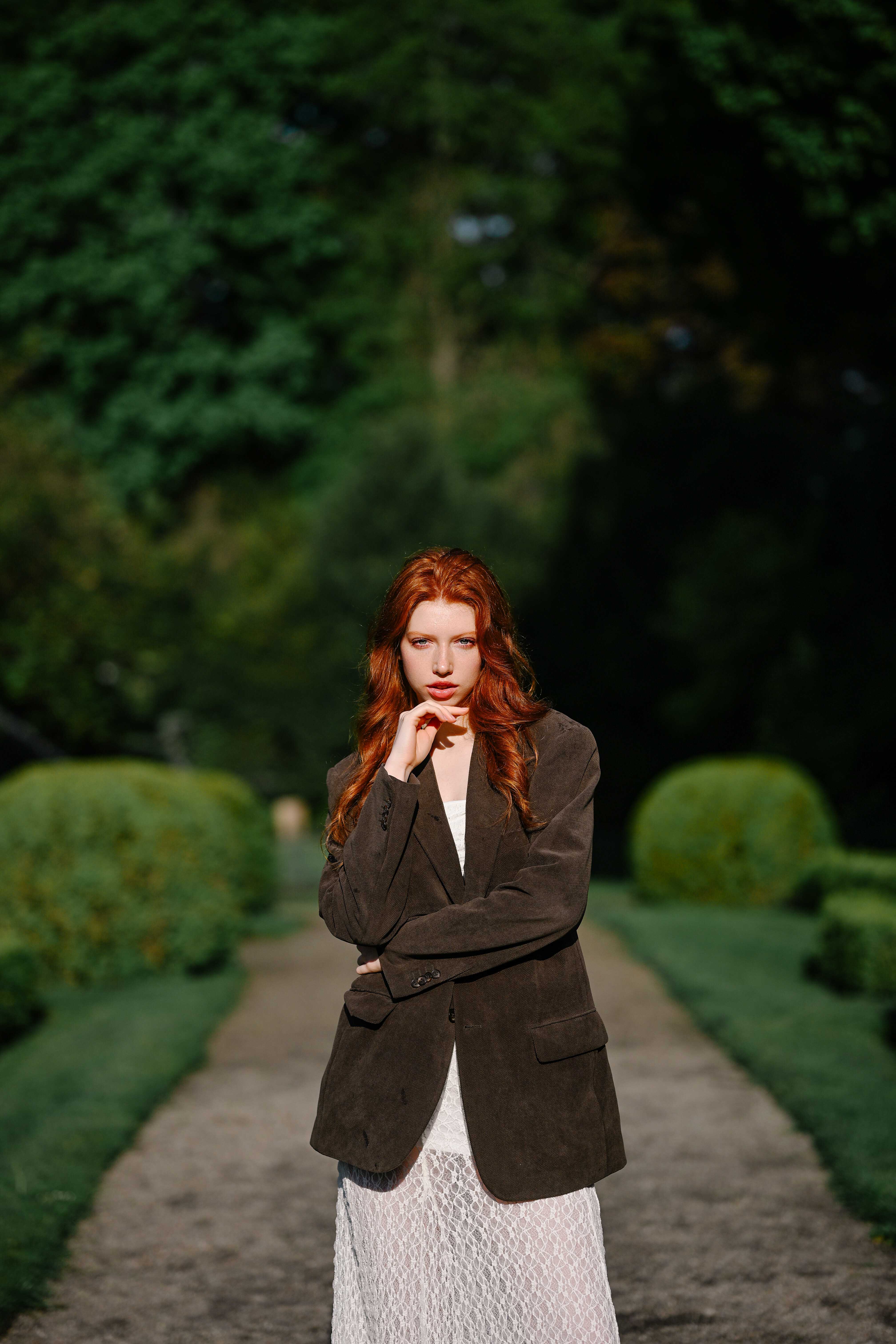 A red-haired model surrounded by lush greenery in Berlin’s Humboldthain park, posing in a brown blazer and white lace dress. Shot by Alina Rudya on a Nikon Z6 III.