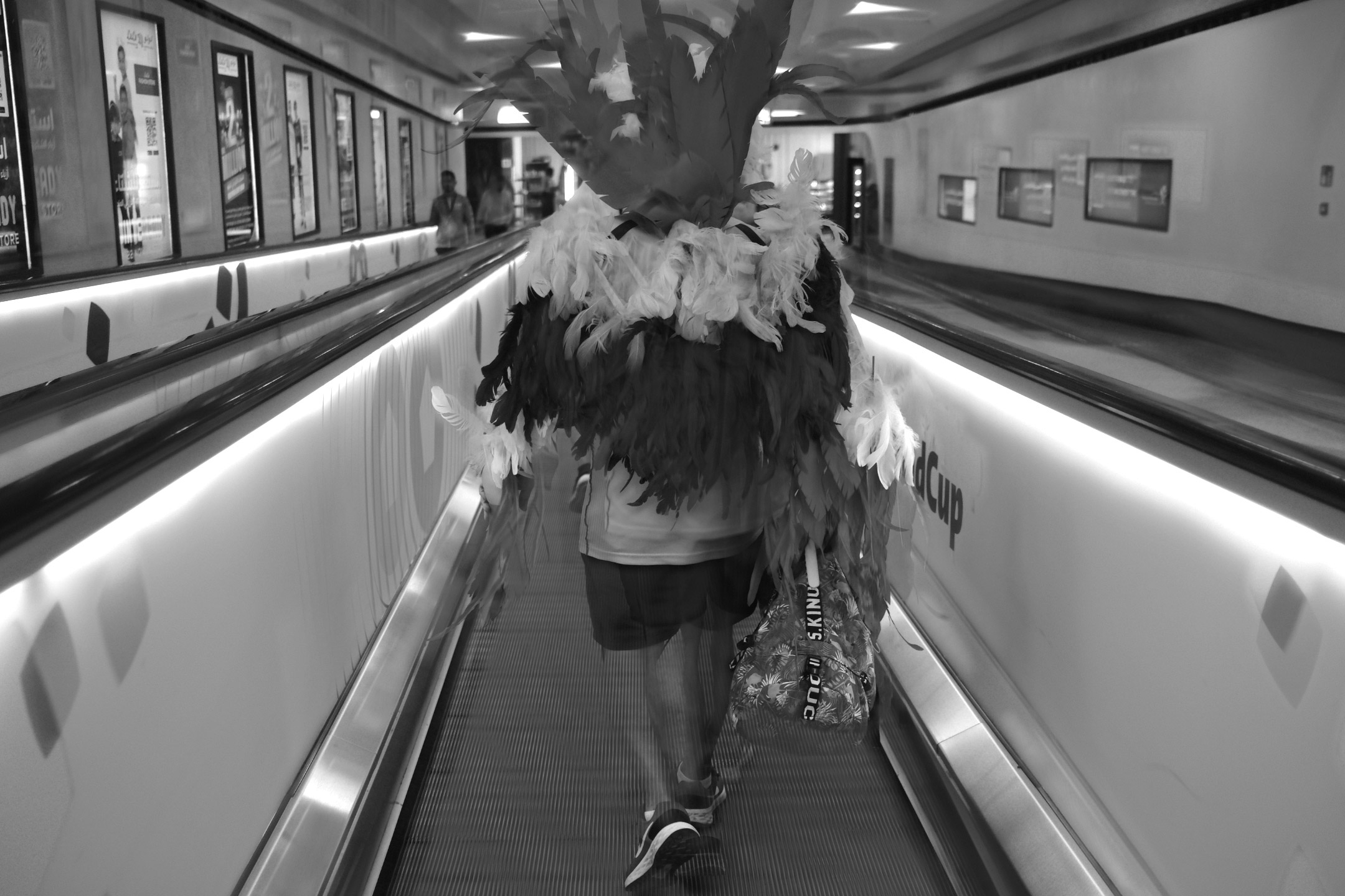 Ein Schwarz-Weiß-Foto eines Fußballfans in einem Federkostüm, der während der Fußball-Weltmeisterschaft 2022 eine Rolltreppe hinunterfährt, aufgenommen von Ryu Voelkel mit einer Fujifilm X100V.