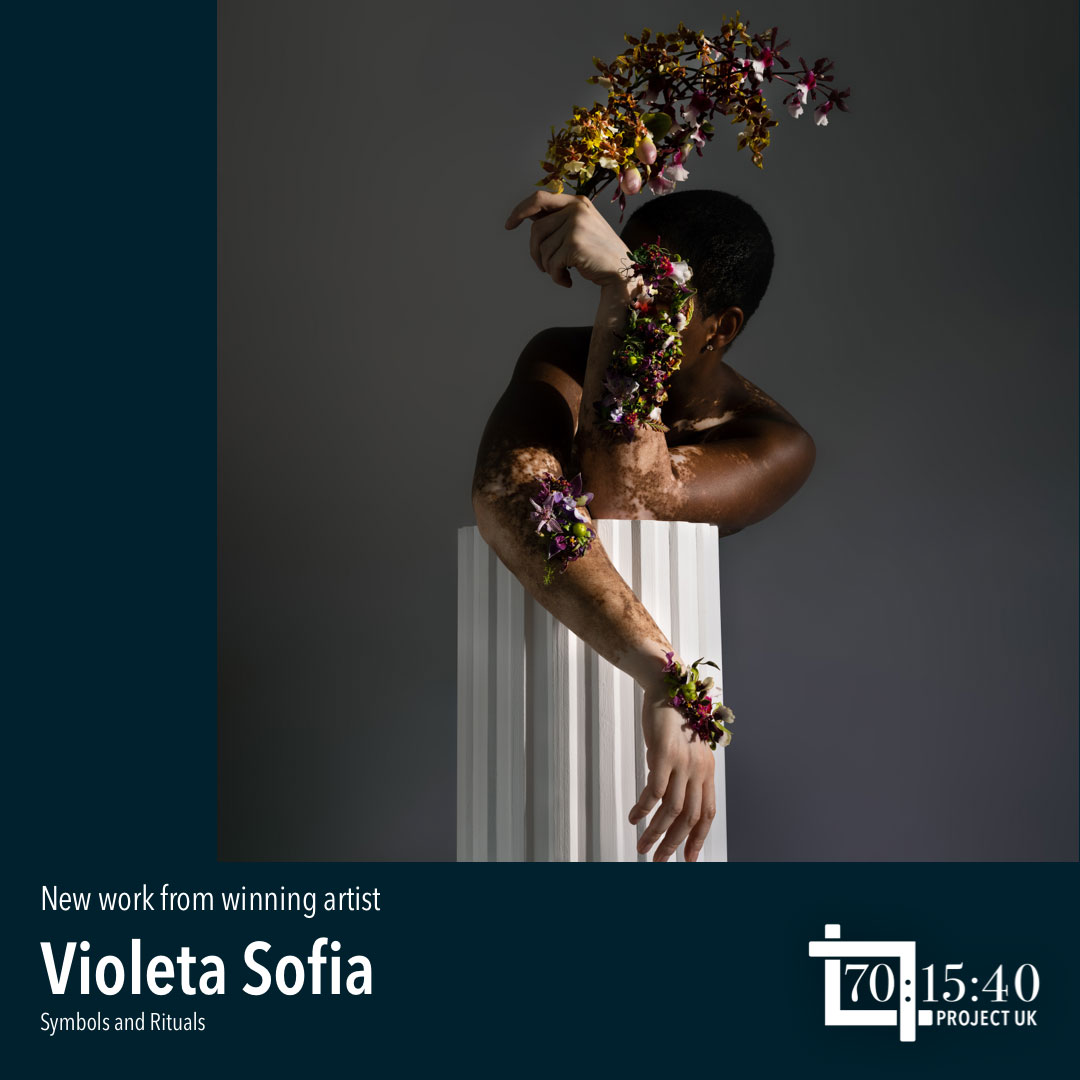 A model holding flowers while leaning on a table covered by white fabric, photo by Violeta Sofia