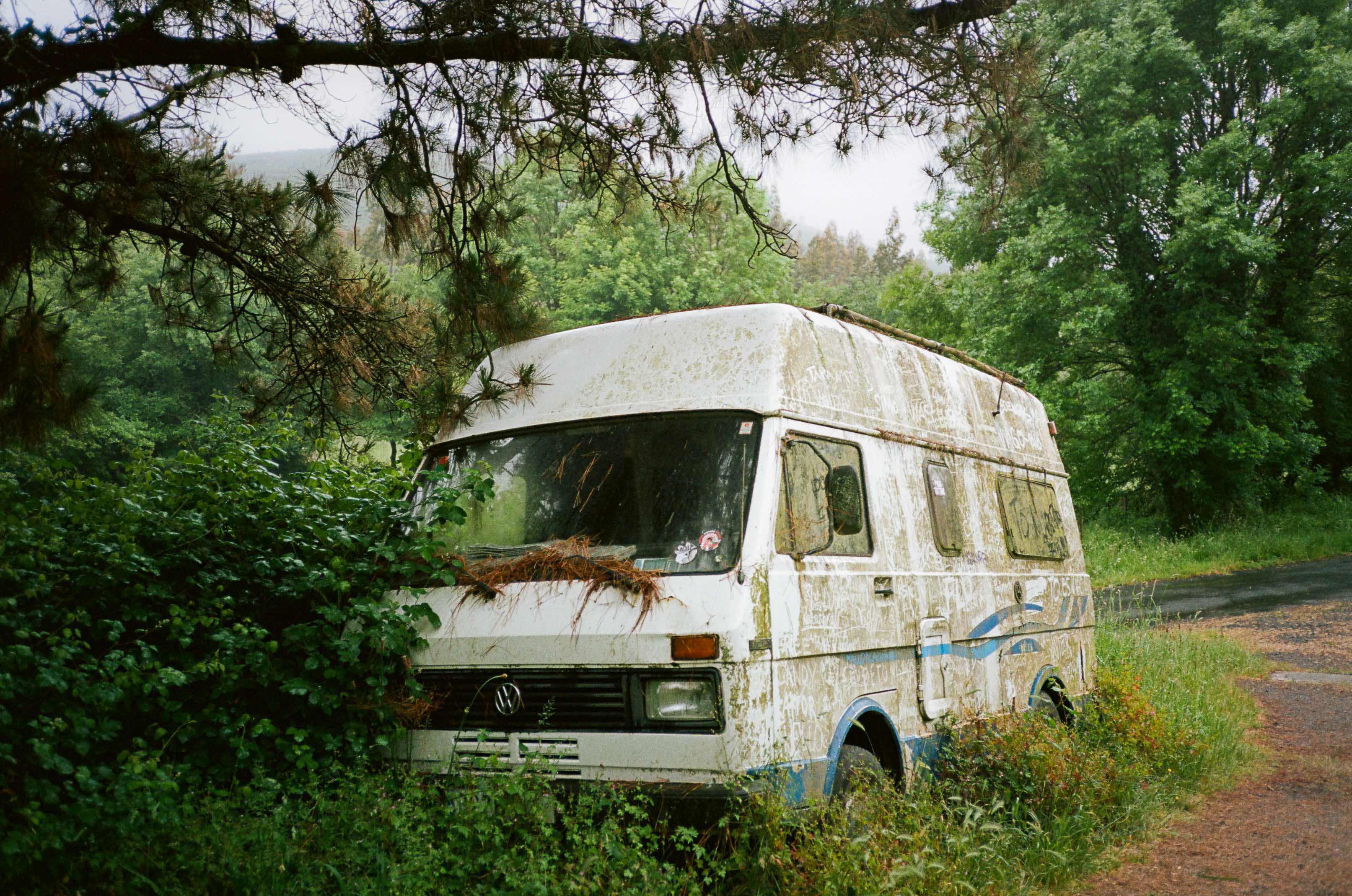 An old blue and white van is abandoned on the side of the road and covered in moss and dirt. By Connor Redmond.