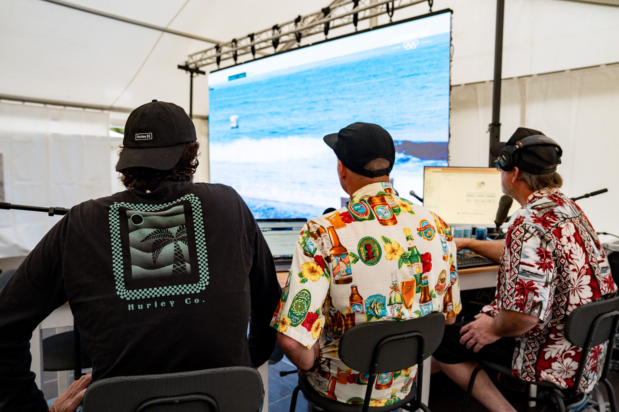Trois hommes en chemise et t-shirt se tiennent assis devant un écran sur lequel se déroule la compétition de surf. Photo prise avec le Sony A9 III par Paul Lavoine.

