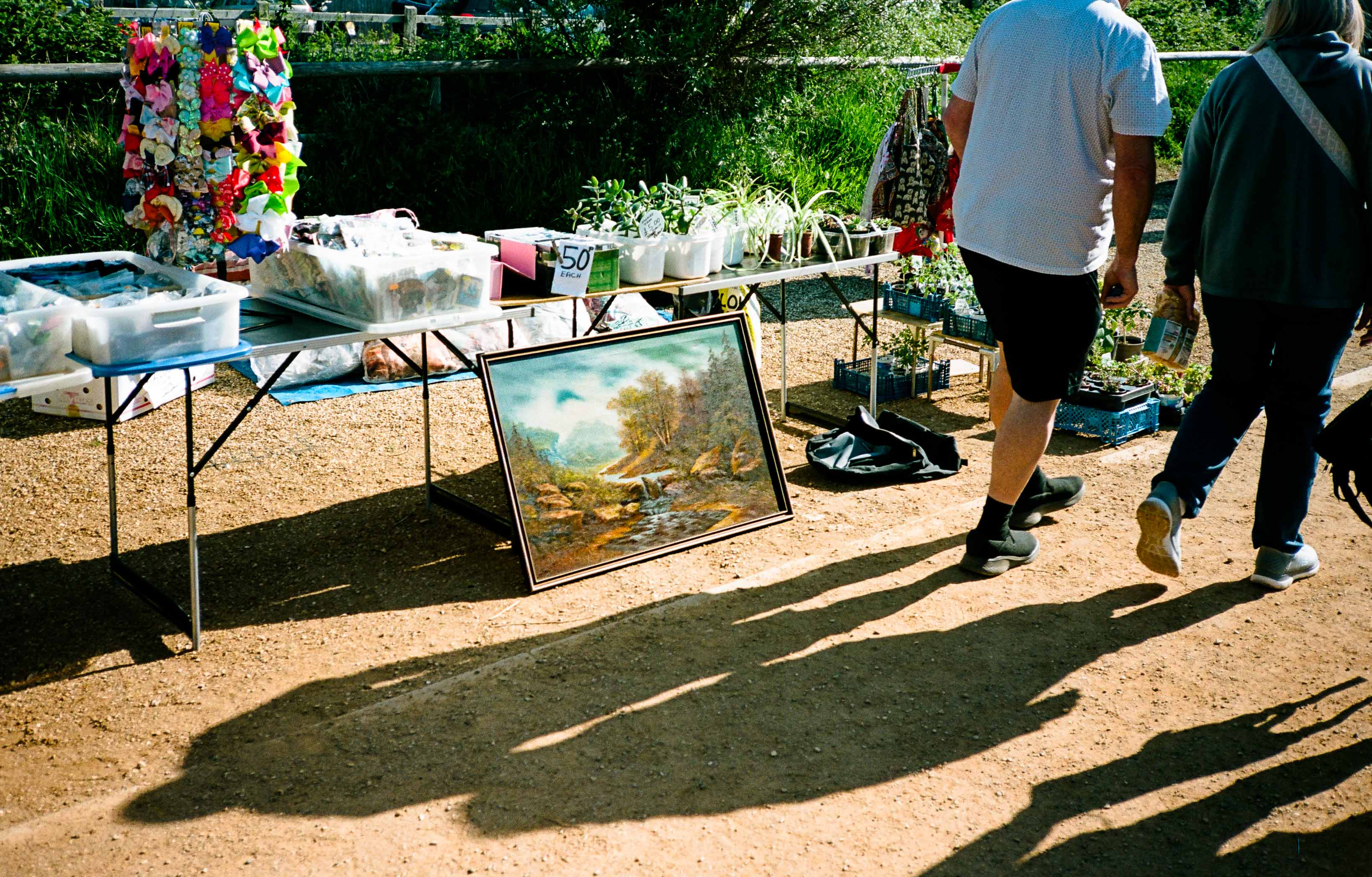 Long shadows of people cast on the floor at a car boot sale, a garish painting sits in the middle of the frame. By Connor Redmond.
