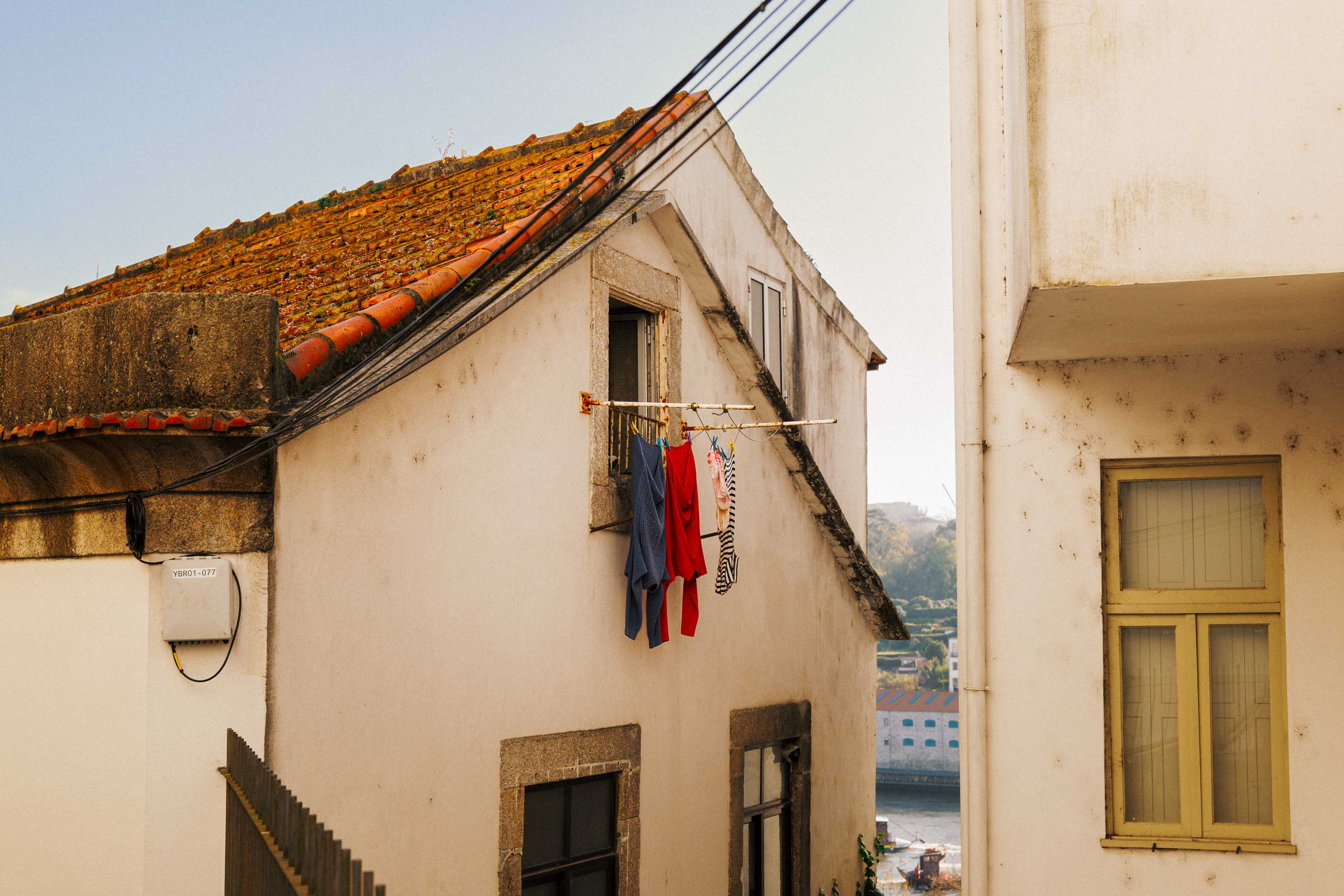  Laundry hanging from a small balcony between whitewashed Porto buildings, with terracotta roof tiles and the river visible in the distance.
