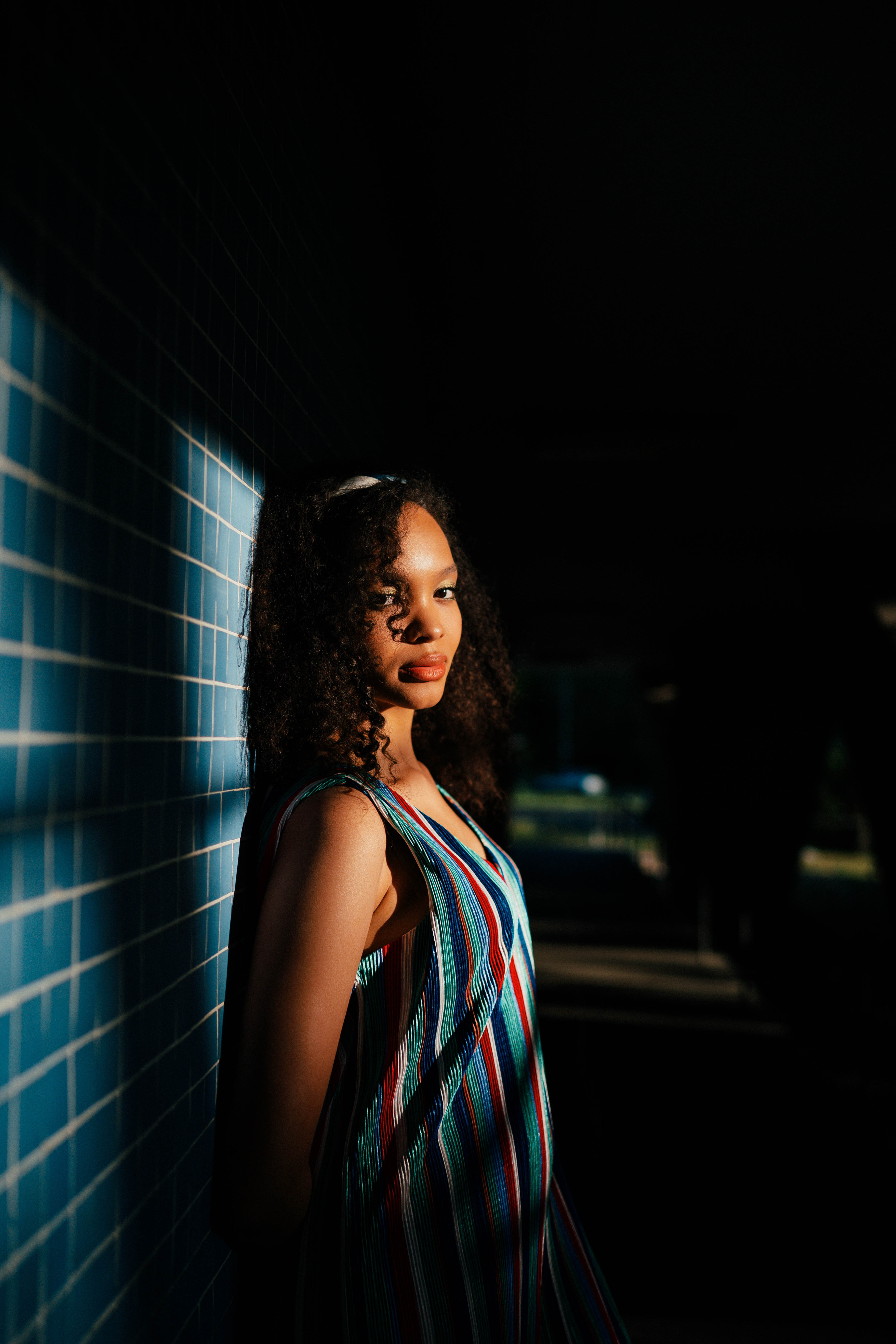 Model in a colorful striped dress, leaning against a blue-tiled wall in Berlin’s Hansaviertel. Sunlight and shadows create a dramatic effect. Shot by Alina Rudya on a Nikon Z6 III.