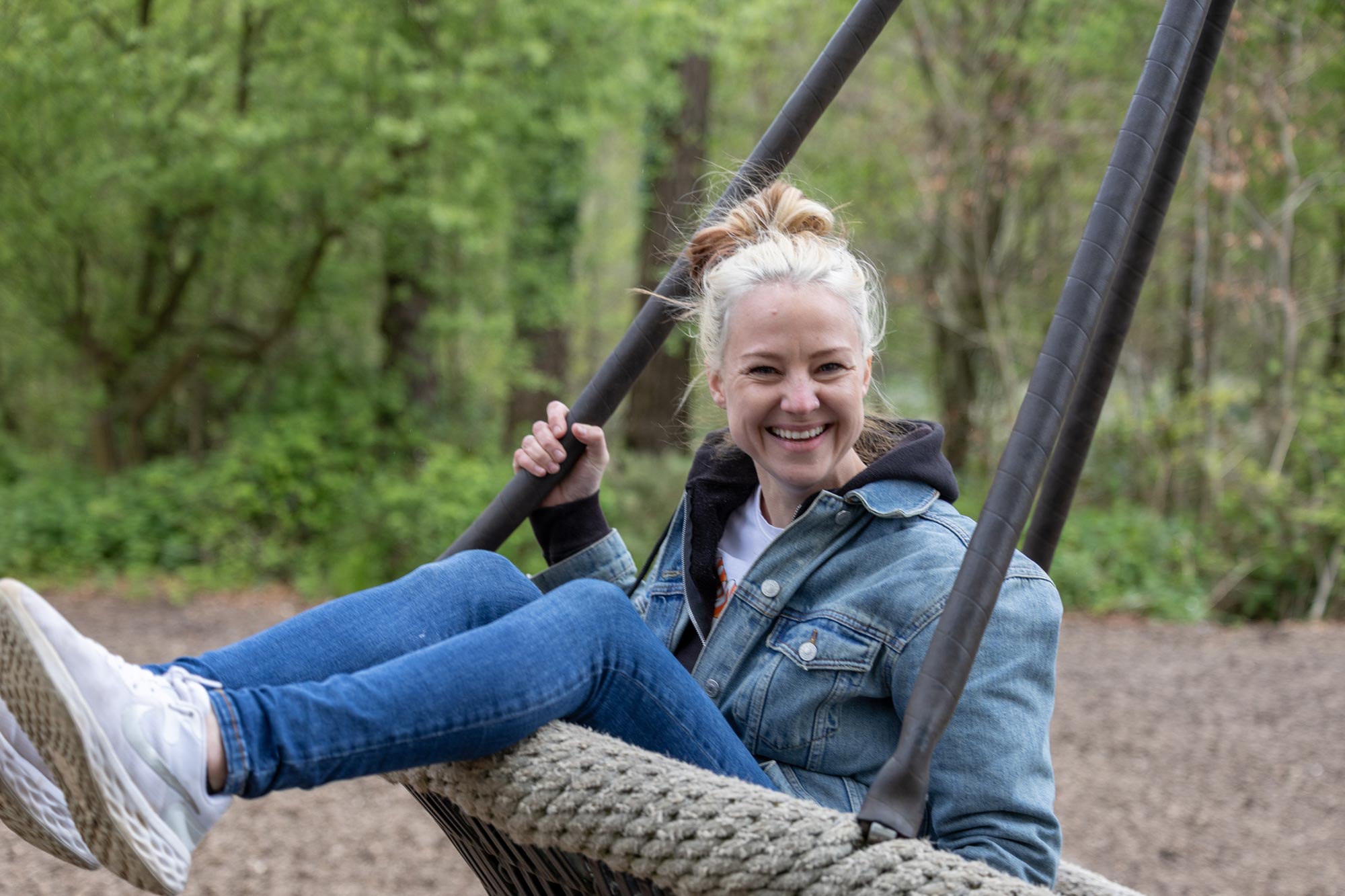 A person smiles on a playground swing, staring straight into camera. The woods are visible in the background.