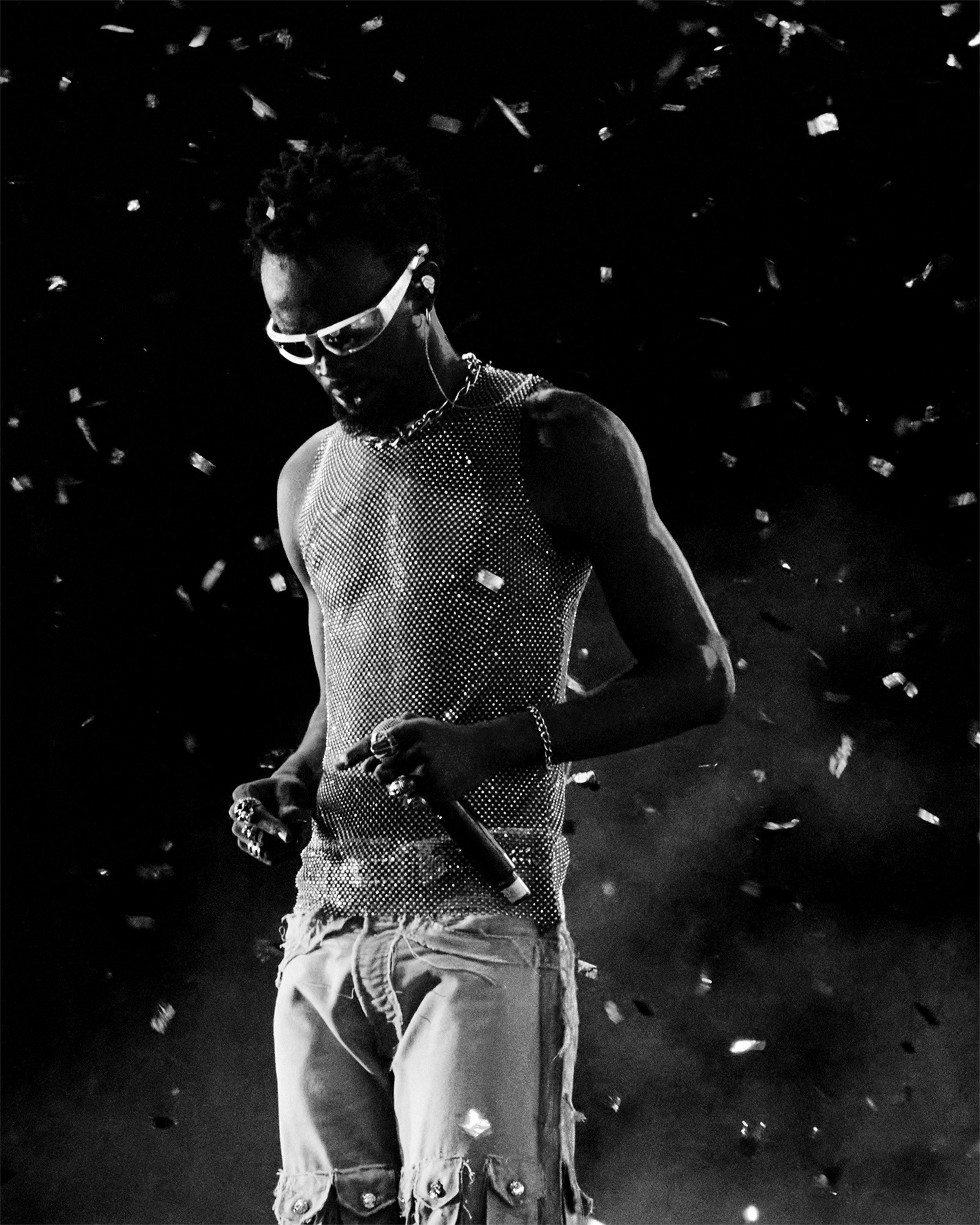 Black and white photo of a Black performer wearing sunglasses and holding a microphone in his left hand, confetti around him, shot by Lisa Marie Asubonteng