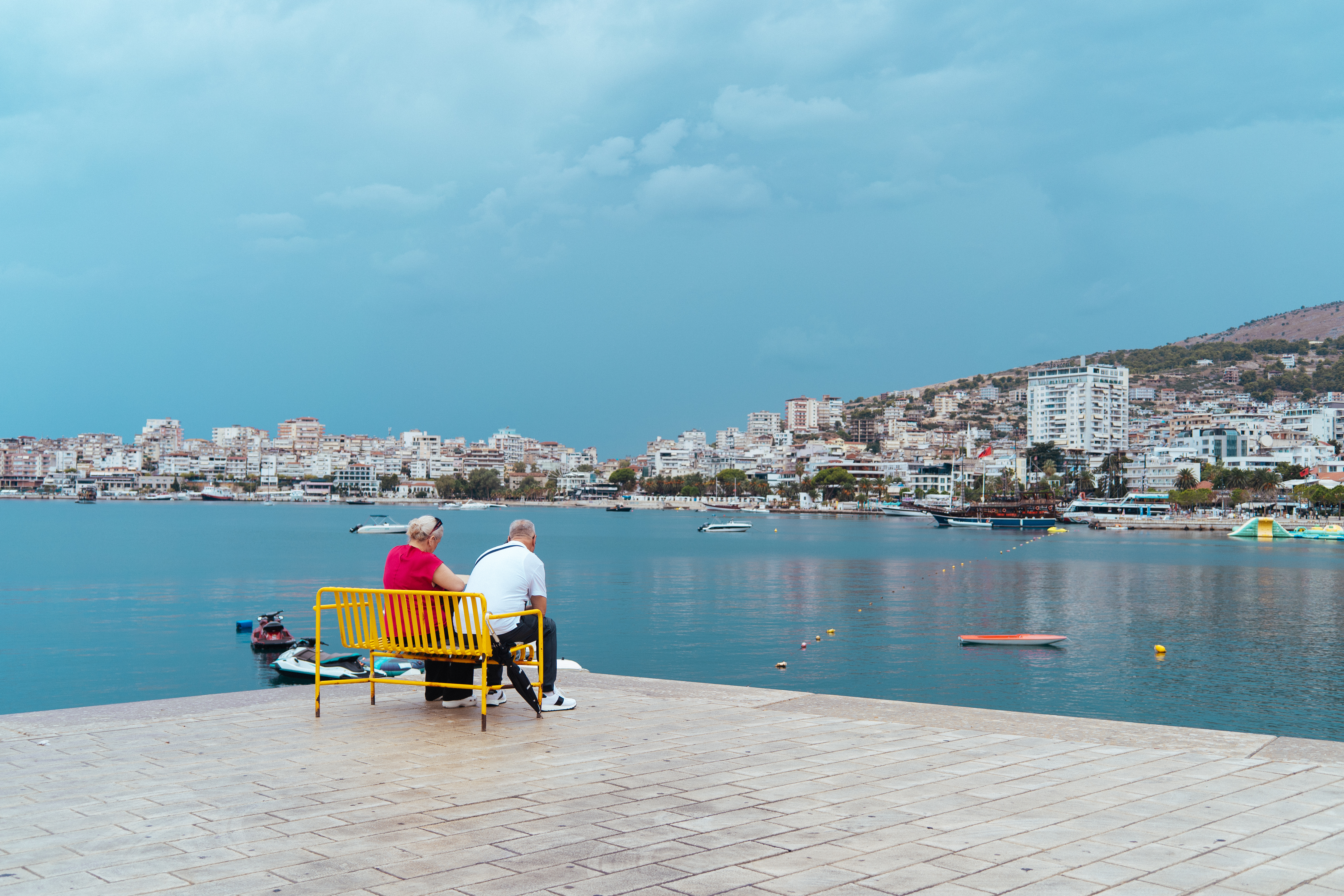 A couple sits on a bright yellow bench overlooking a coastal city, as storm clouds gather across the horizon.