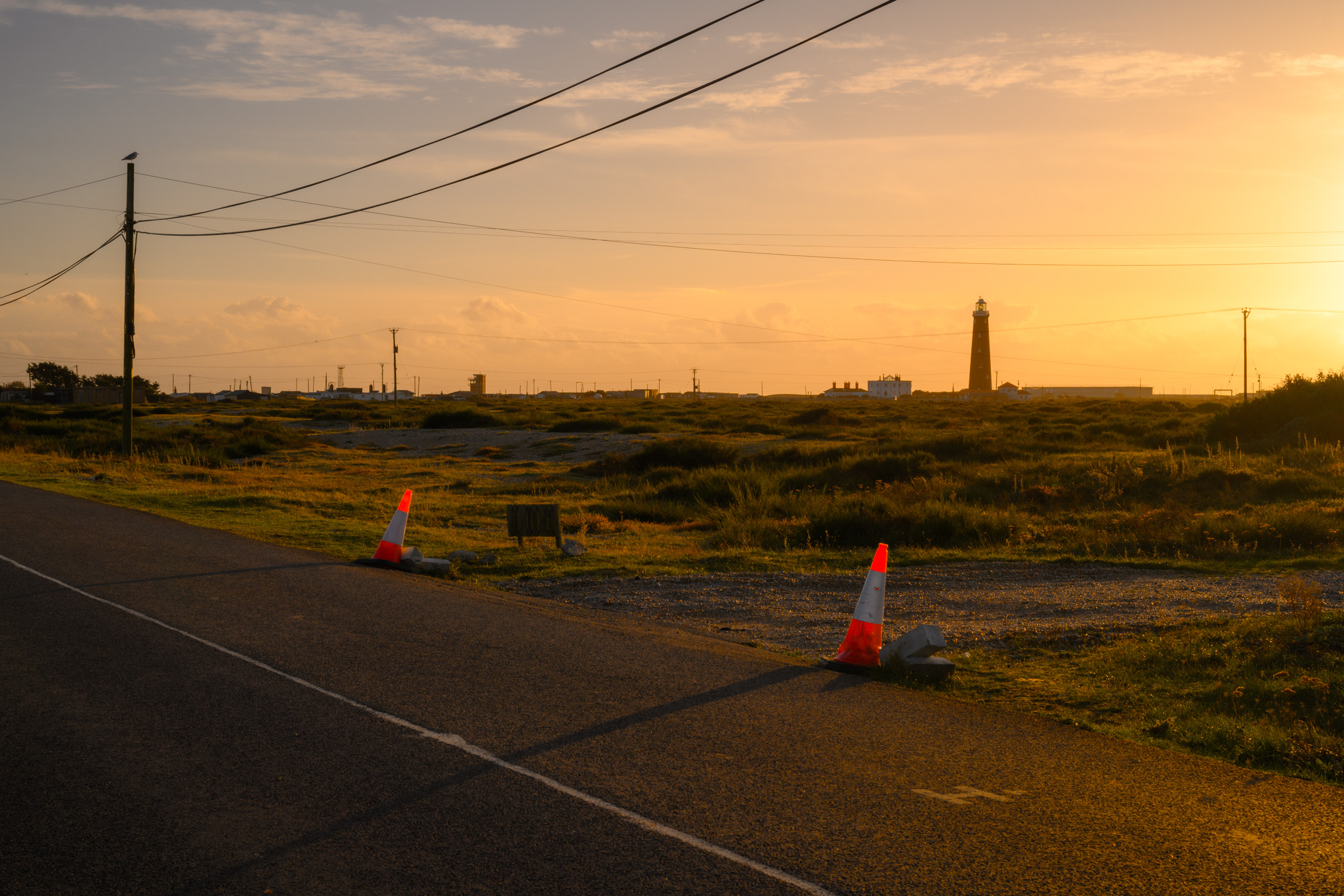 Zwei Straßenkegel im Vordergrund sind beleuchtet und werfen lange Schatten bei Sonnenuntergang am Strand von Dungeness, Kent. Im Hintergrund sind Strommasten, Drähte und ein Leuchtturm zu sehen. Von Connor Redmond.
