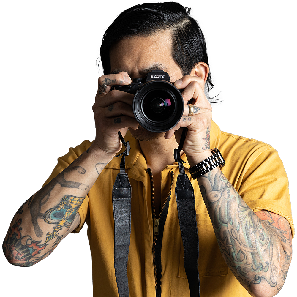 Een getatoeëerde fotograaf, gekleed in een geel shirt en met een horloge om, richt een Sony-camera op de kijker.
