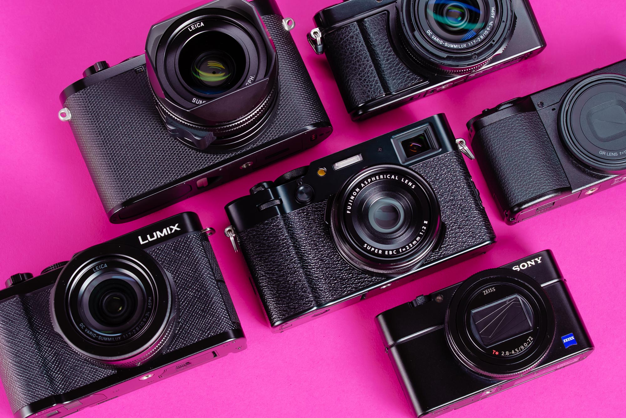 A selection of used everyday carry compact cameras on a pink background. From L-R: a Panasonic Lumix L10, Fujifilm X100VI, Sony RX100 VII and Ricoh GR IV.