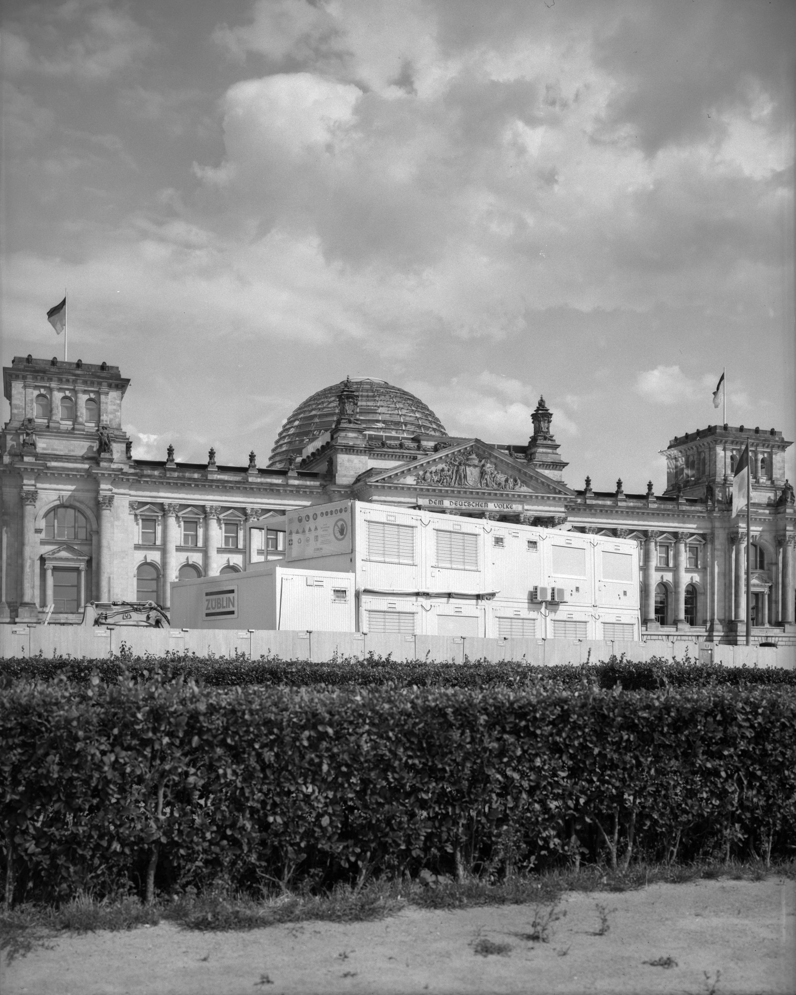Black-and-white film photograph of Berlin’s Reichstag building under cloudy skies, with construction containers and manicured hedges in the foreground. Shot by Austin Fassino on 35mm film.
