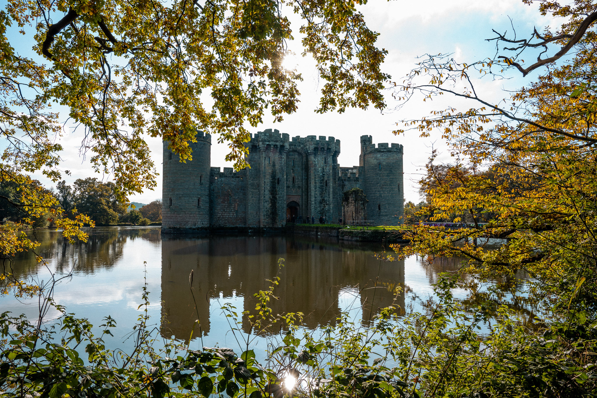 Bodiam Castle surrounded by a moat, reflected in calm water and framed by autumn trees, photographed with the Nikon Zf by Jakub Golis. 
