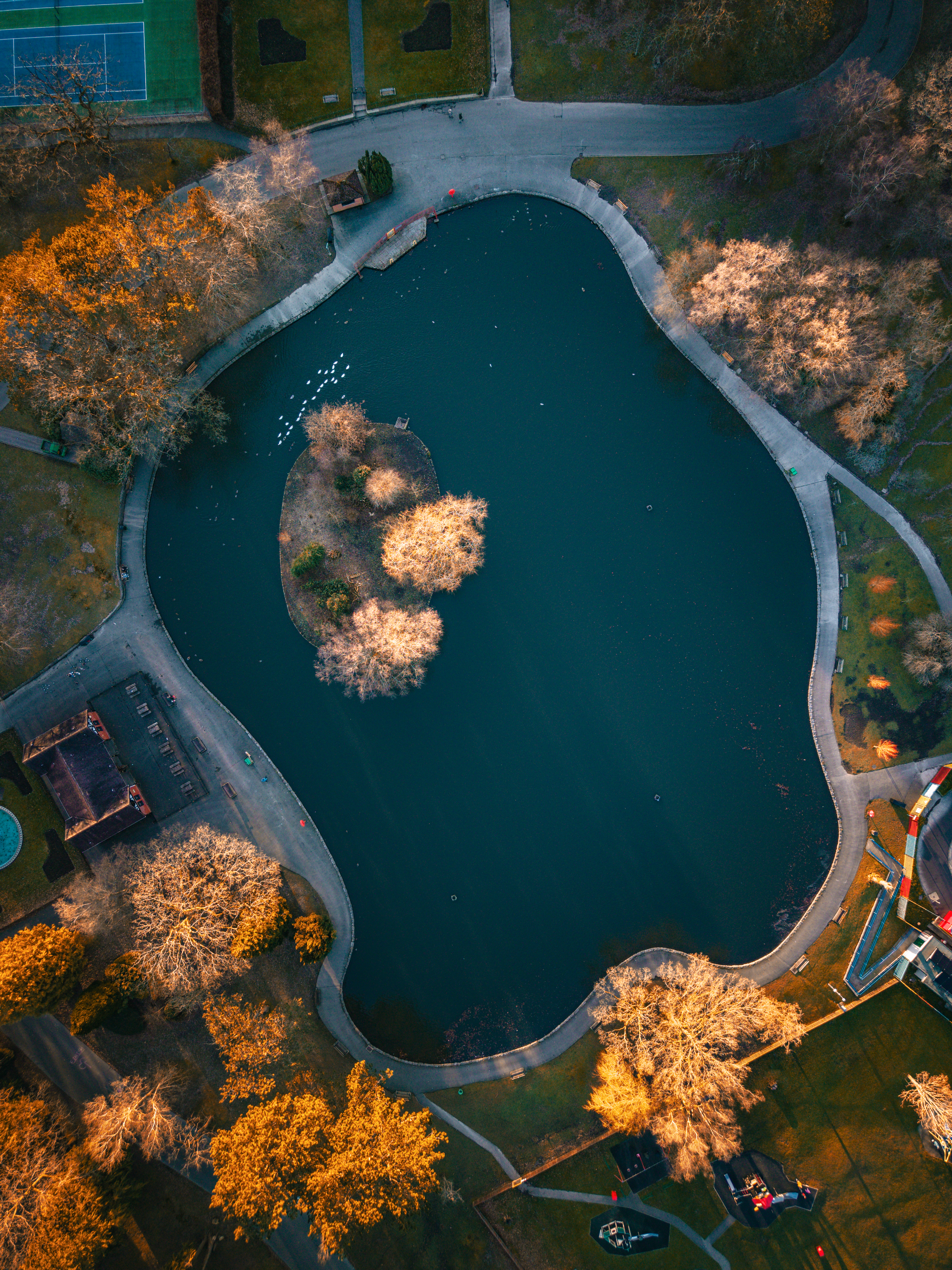 Imagem aérea captada com o DJI Mini 5 Pro, mostrando um lago urbano rodeado por caminhos, árvores e zonas verdes. A luz quente do fim do dia destaca as copas das árvores e cria contraste com o azul profundo da água.