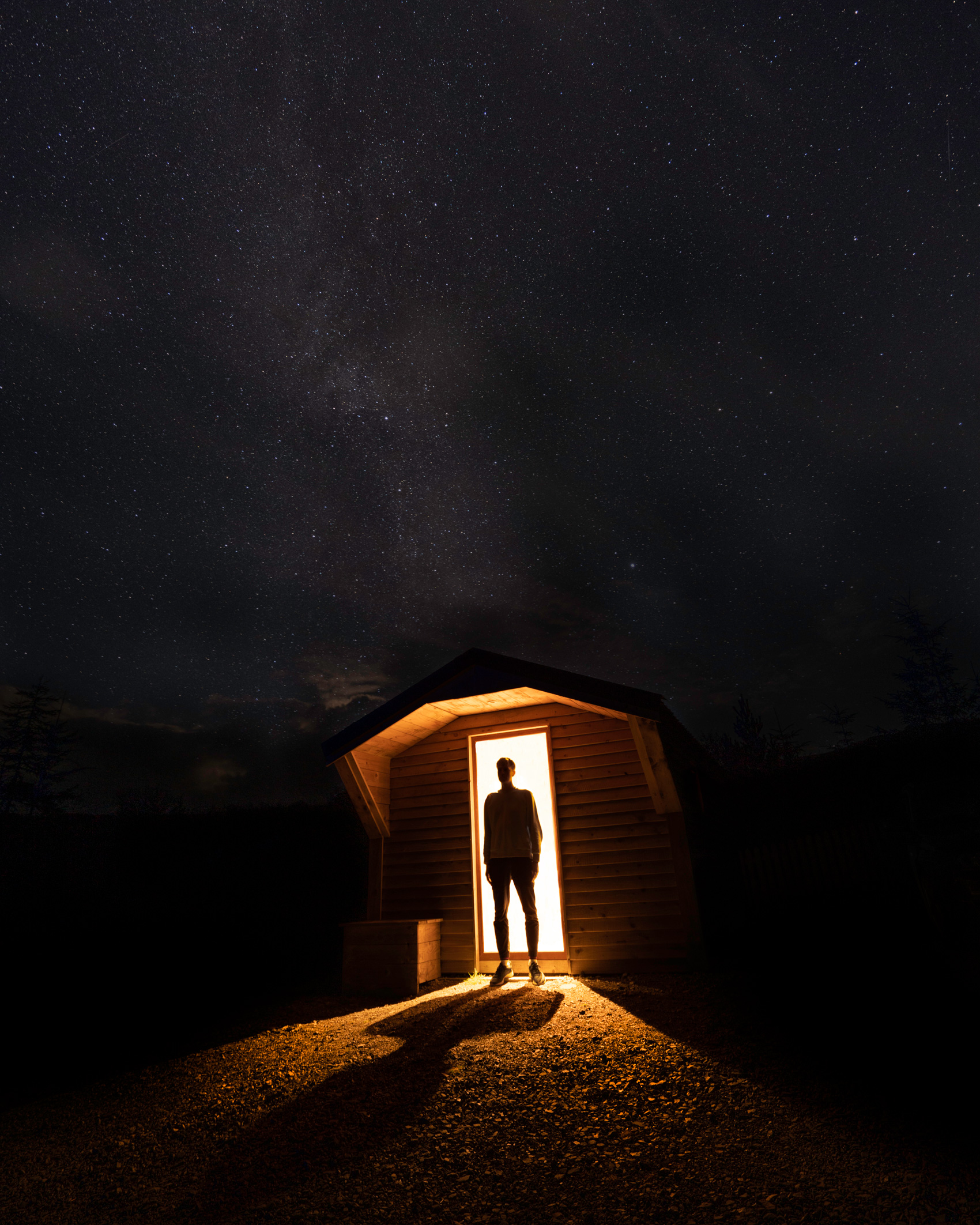 A long exposure of the silhouette of a person standing in the bright doorway of a cabin on the Isle of Skye, with the Milky Way overhead, shot by Jordan Lancaster-Graham with a Sony A7R III