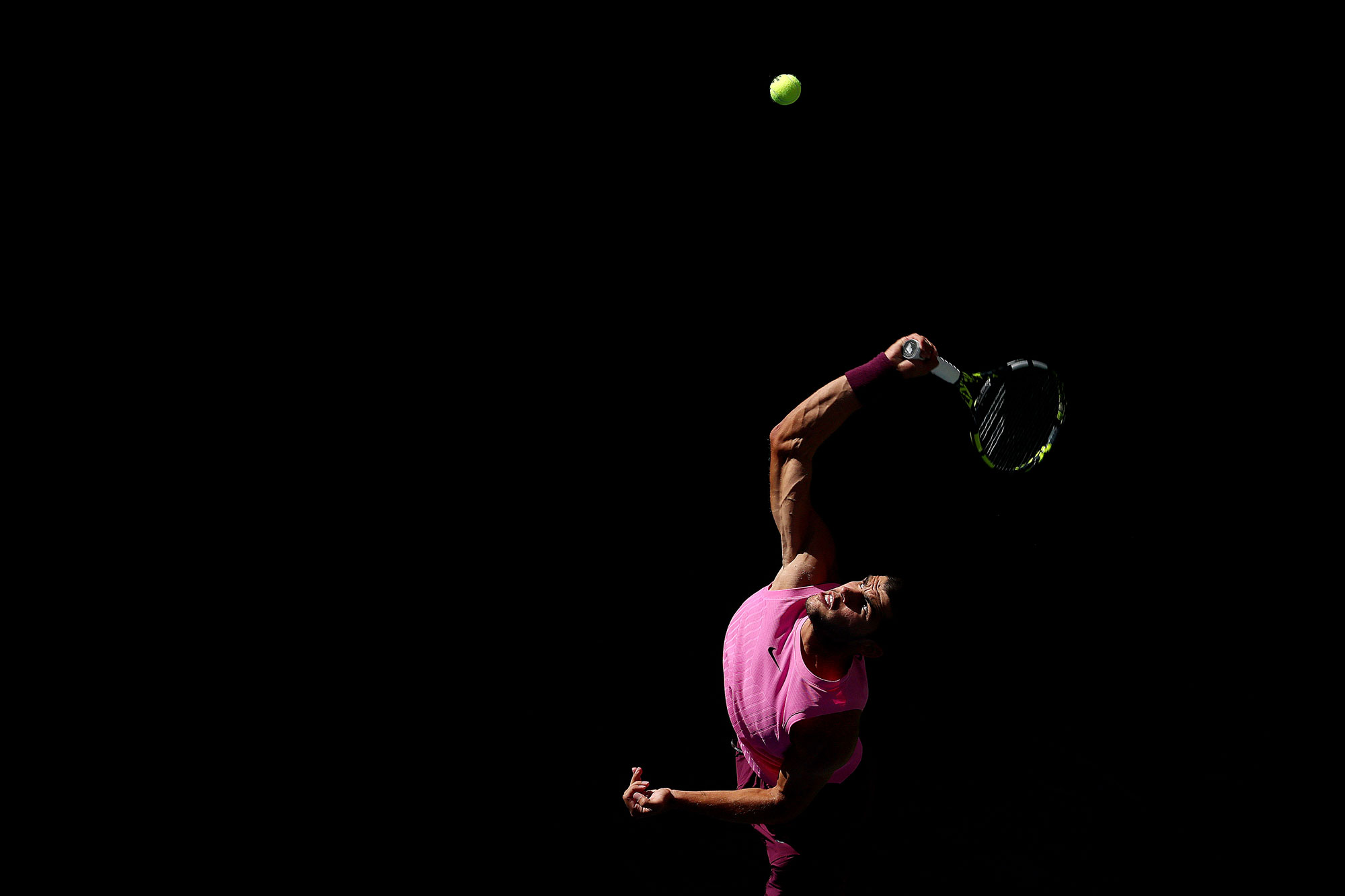 Carlos Alcaraz of Spain serves, his right arm stretched back. The tennis ball is illuminated and mid-air, with the background of the shot completely pitch black.