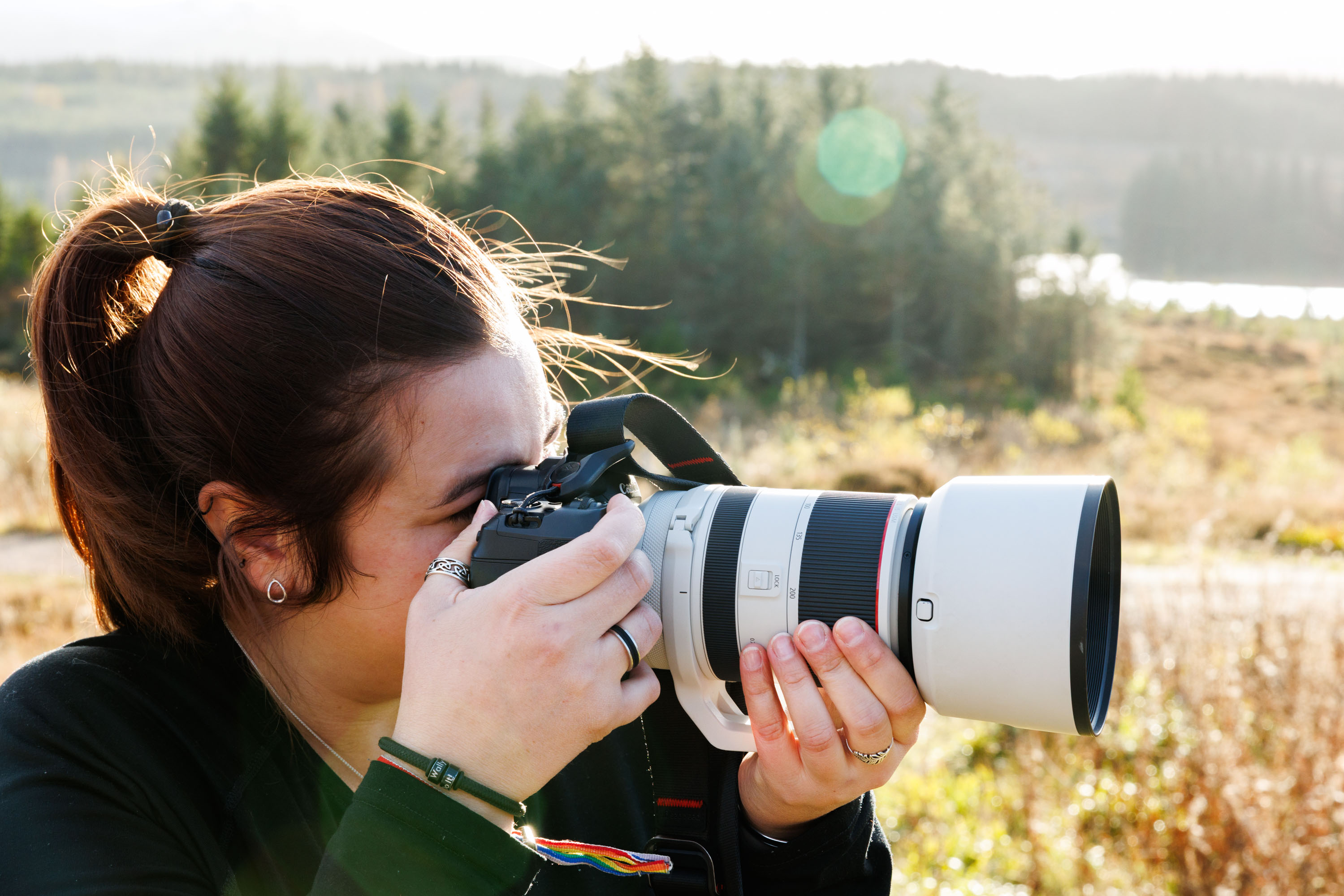 Person photographing an outdoor scene with a Canon camera and a white telephoto lens fitted with a lens hood
