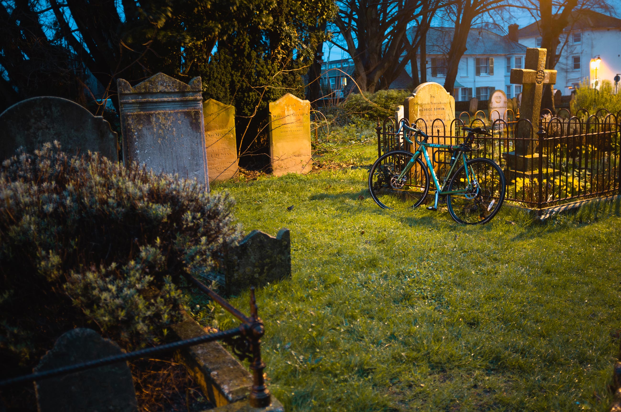 A mountain bike is propped up against some railings in a graveyard at dusk, photo by Jakub Golis with Fujifilm X100