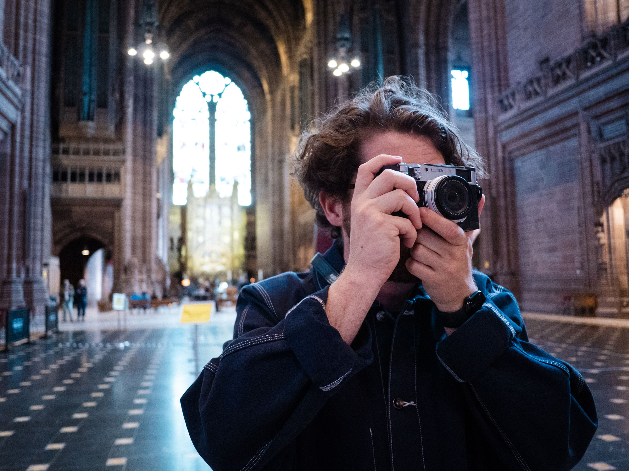 Person, die in einer Kirche mit einer Kamera ein Foto macht. Der Hintergrund ist unscharf und Licht strömt durch ein großes Buntglasfenster ins Kirchenschiff.