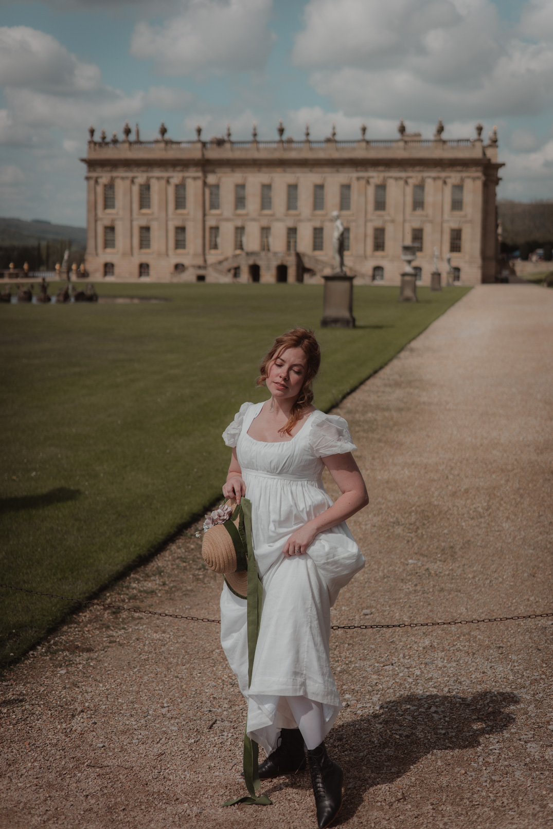 Model wearing white dress holding a hat sitting on a stool in front of flowers at Chatsworth House in Derbyshire, UK, photo by Liza Roberts with a Canon EOS R6
