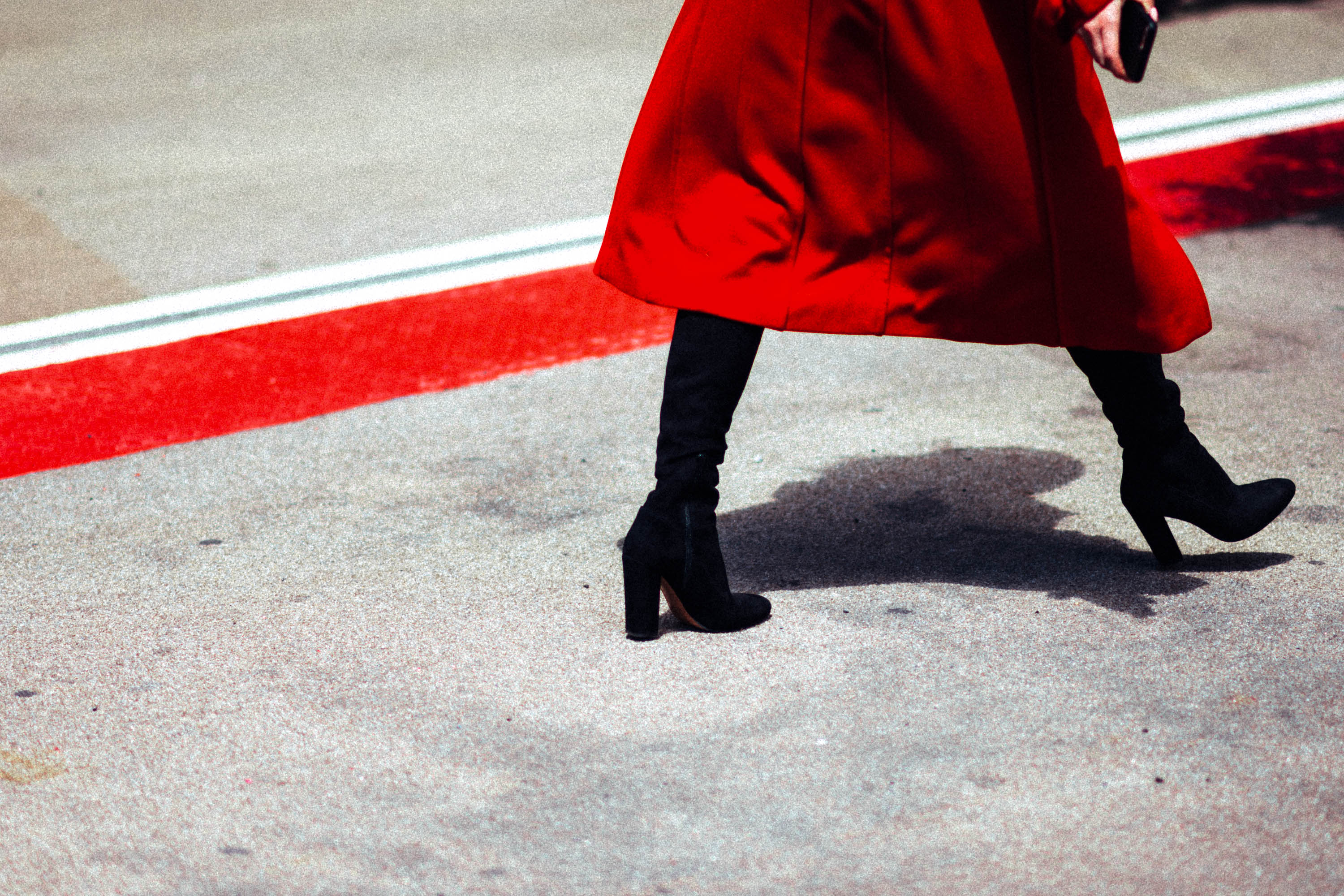 Person wearing red coat and black boots walks on a concrete path alongside a bright red line, street photo by Ian Howorth