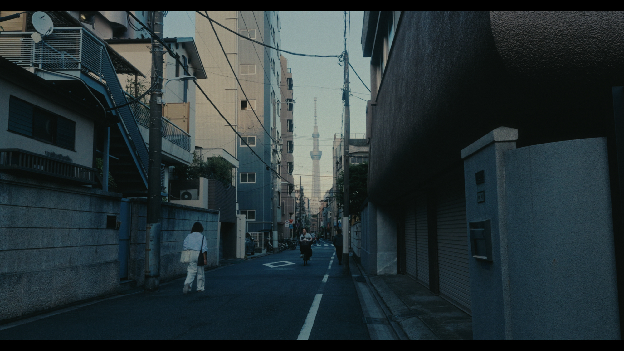 Pedestrians walk down a narrow urban street in Tokyo with a tall tower rising in the distance.