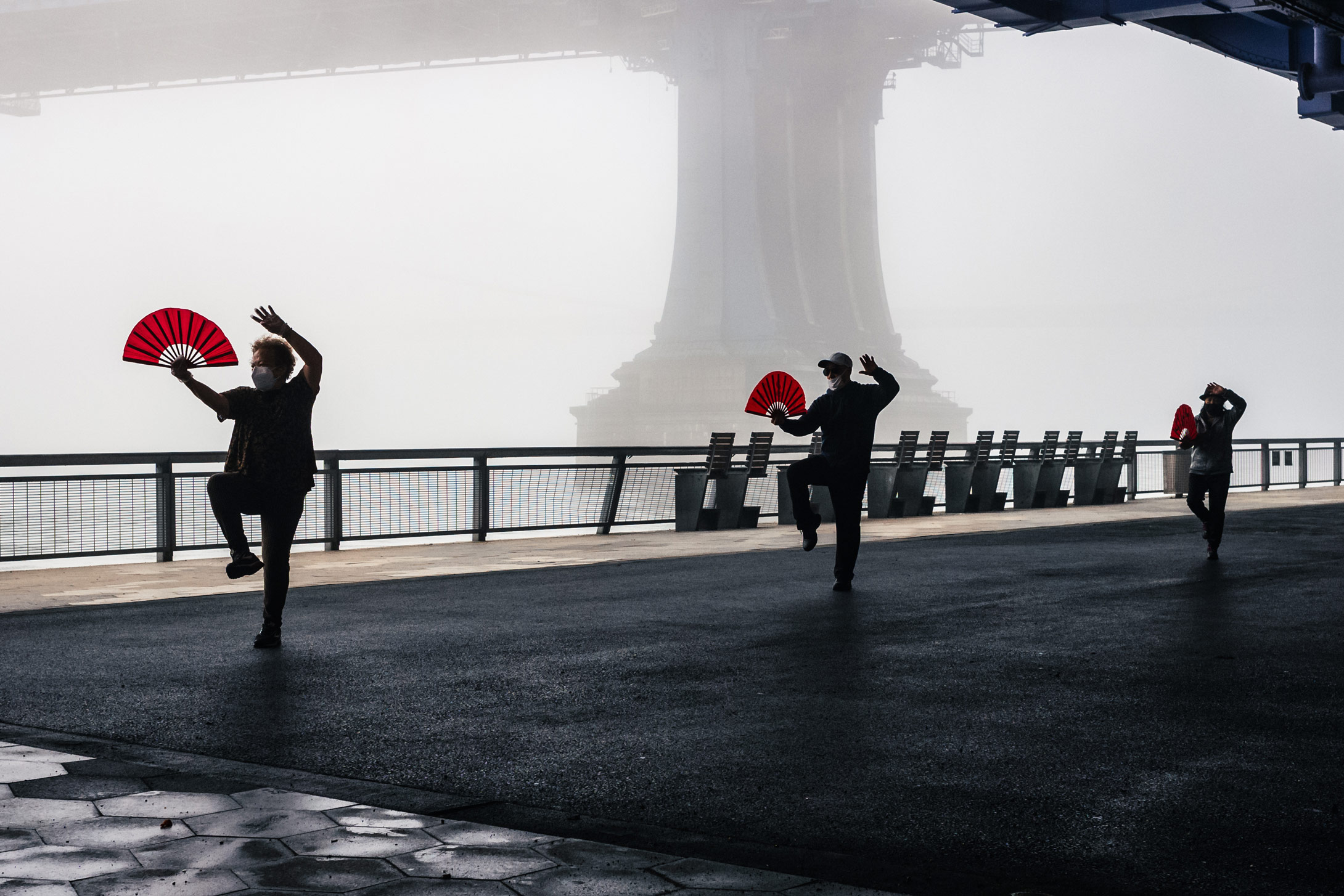 Mensen houden rode waaiers vast en doen een tai chi-houding voor de Brooklyn Bridge op een mistige dag in New York City, gefotografeerd door Manuel Pena.