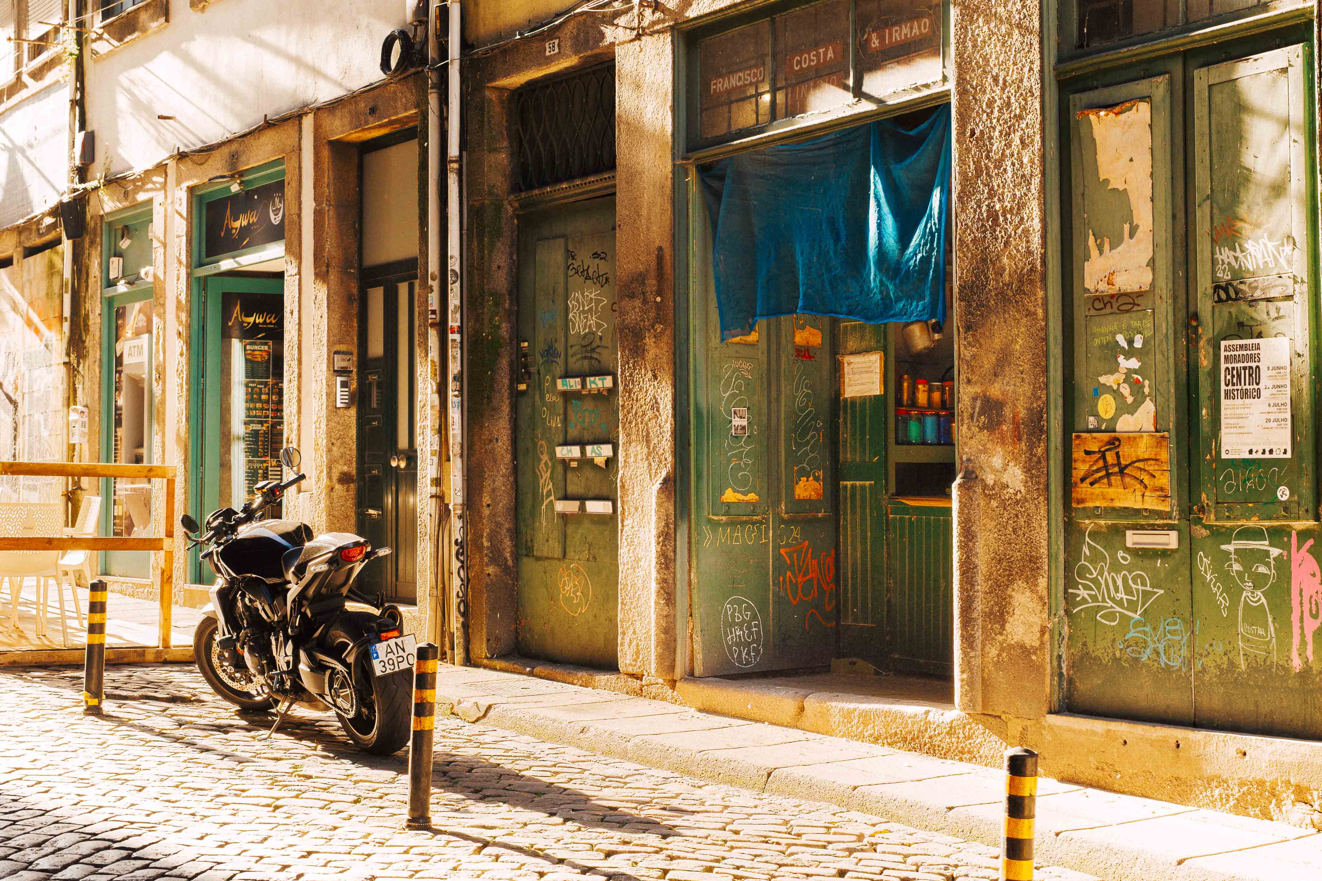 A sunlit Porto street with a parked motorbike in front of weathered shopfronts covered in graffiti and peeling paint.
