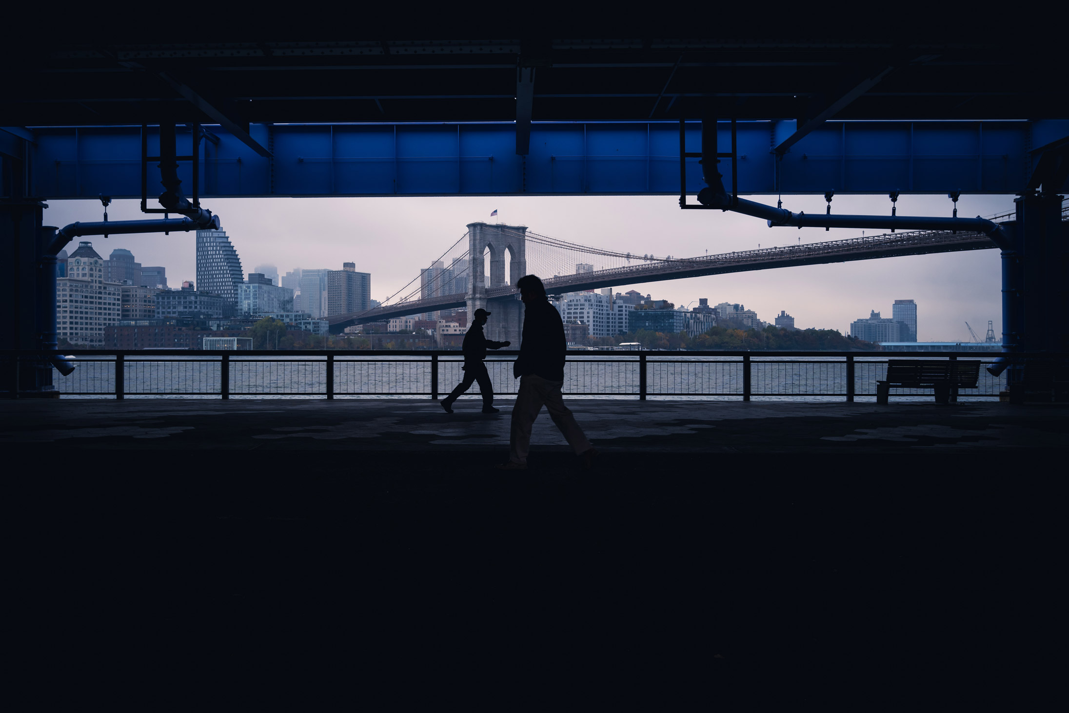 Mensen lopen onder een schaduwrijke loopbrug langs de East River in NYC met de Brooklyn Bridge op de achtergrond, gefotografeerd door Manuel Pena.