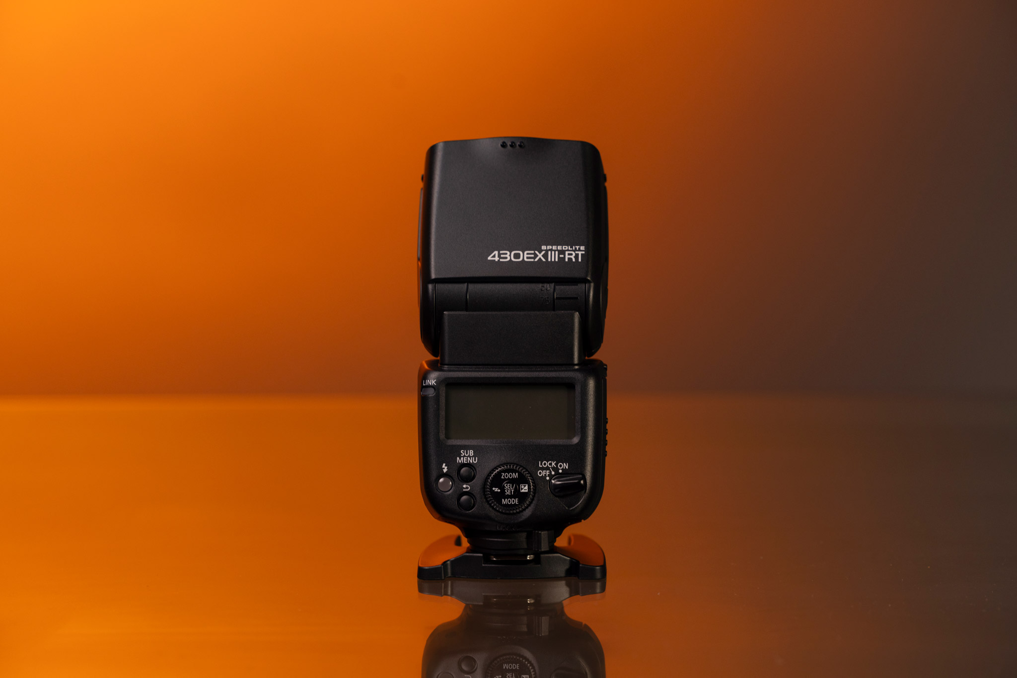Le Canon Speedlite 430EX III-RT en avant-plan, posé contre un fond orange.